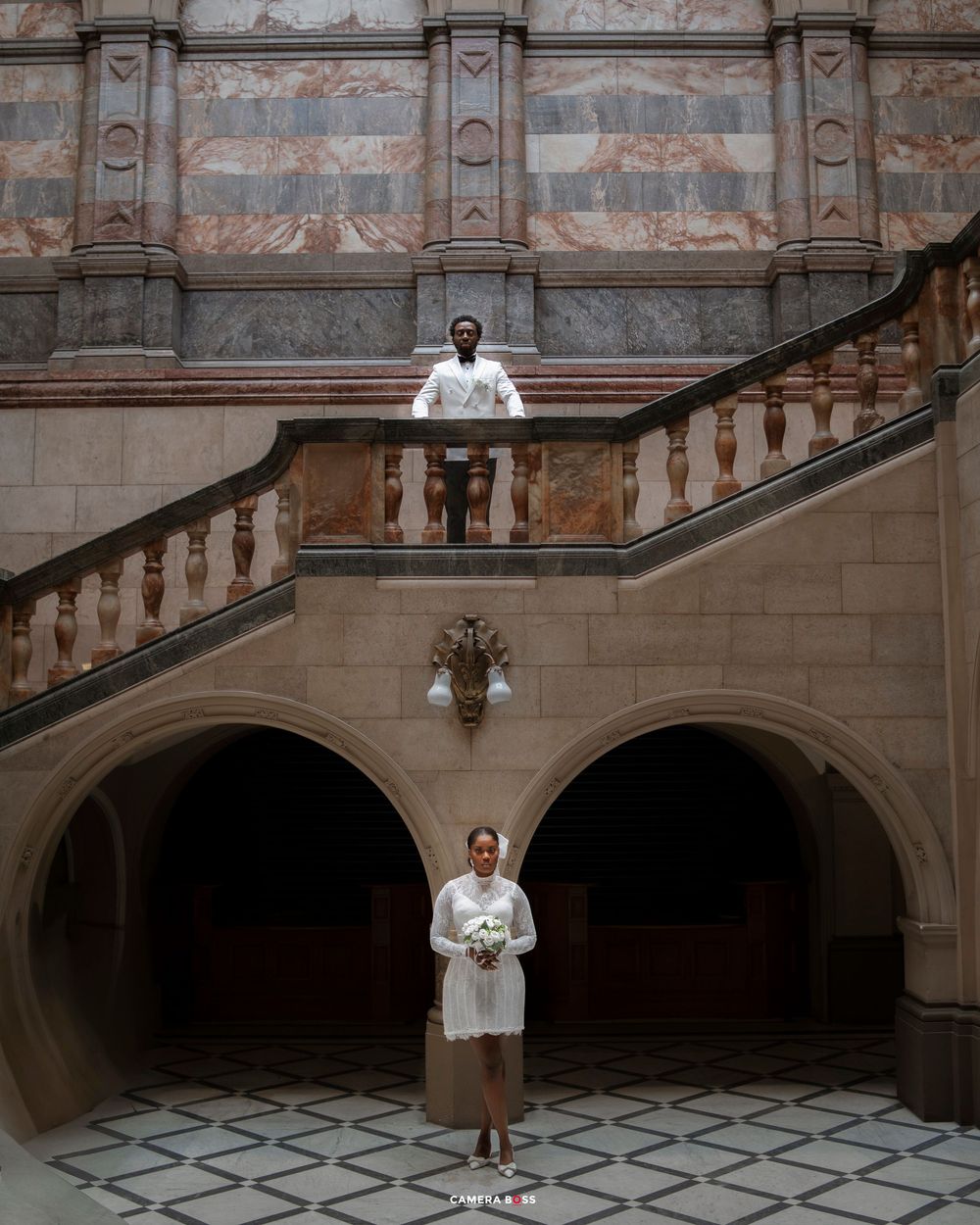 Couple poses on grand marble staircase in ornate historic building, bride holding bouquet below groom on balcony.