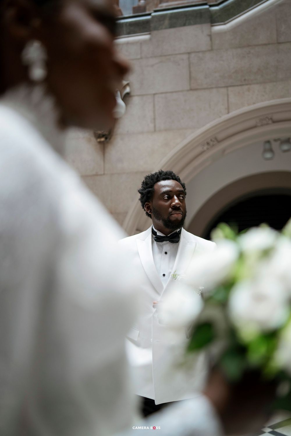 Groom in white tuxedo jacket with bow tie watches bride approach during wedding ceremony in elegant stone venue.
