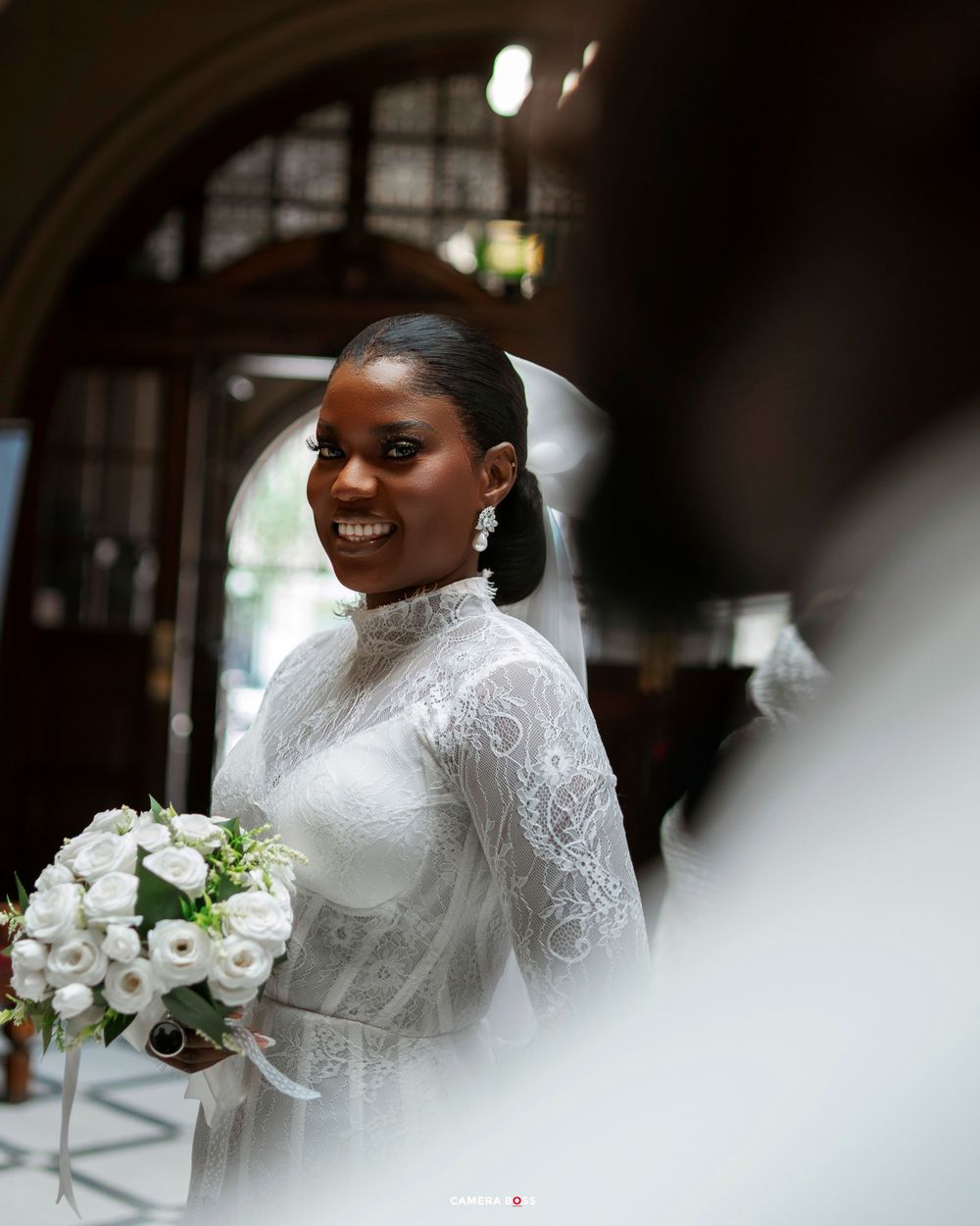 Smiling Black bride in elegant white lace high-neck gown holding white rose bouquet inside a church.