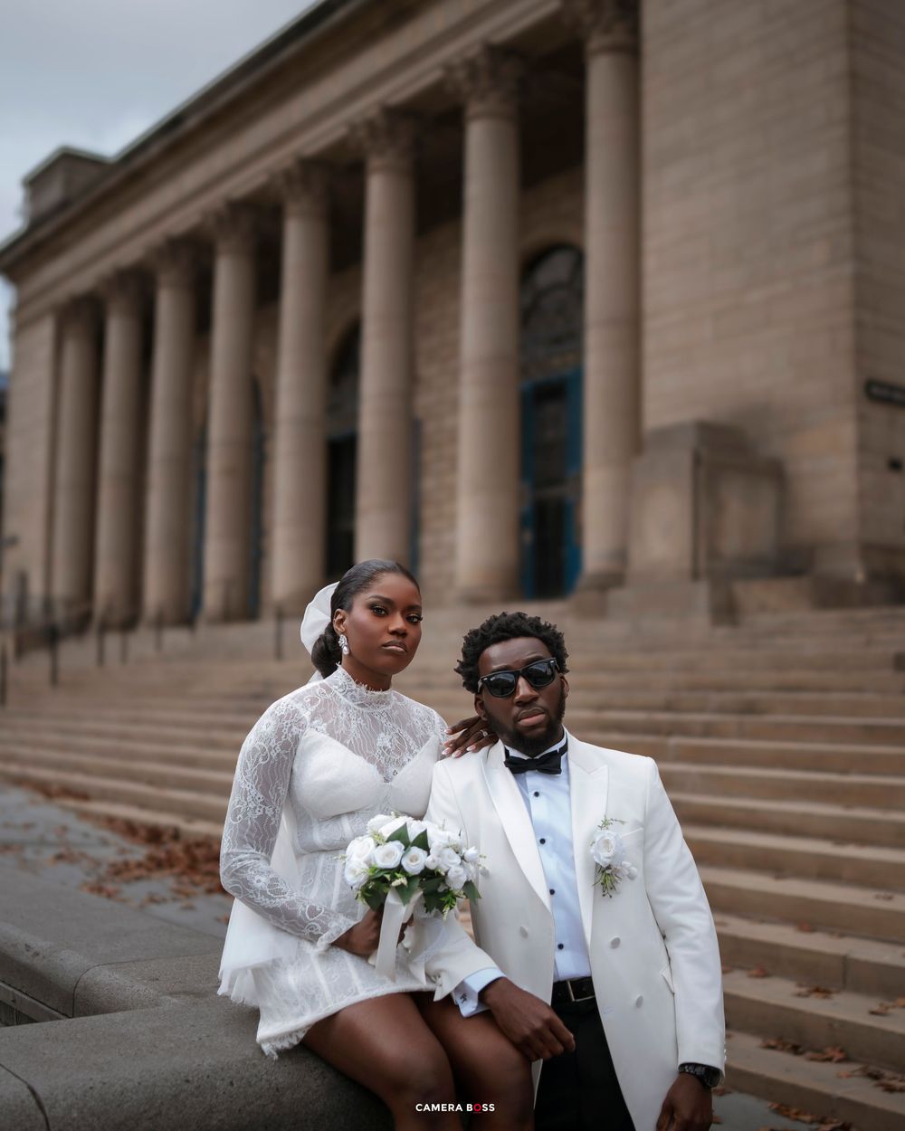Elegant couple in white wedding attire pose on steps of neoclassical building with bouquet.