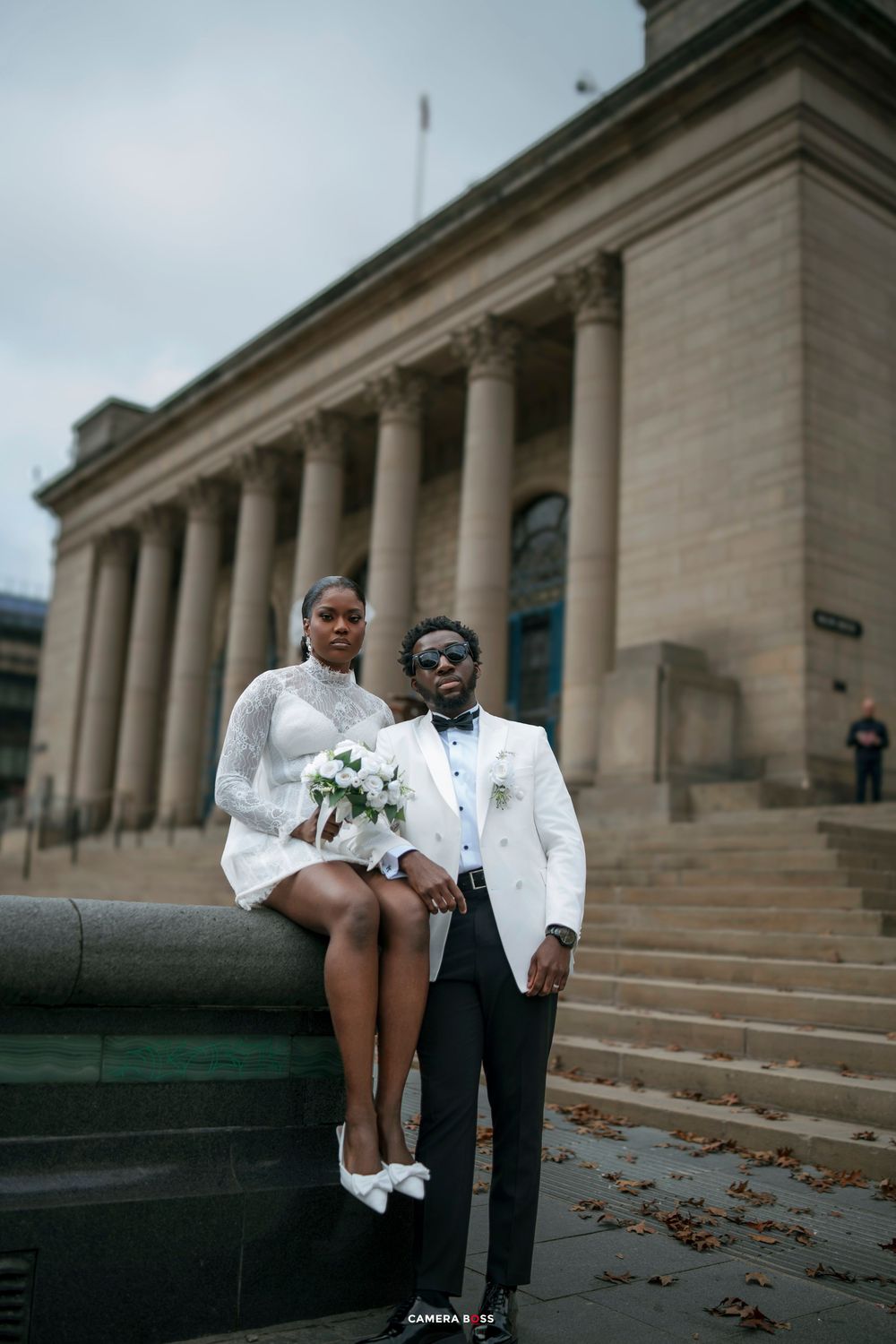 A bride in a white mini dress and groom in a white tuxedo pose at a grand neoclassical building for their wedding photos.