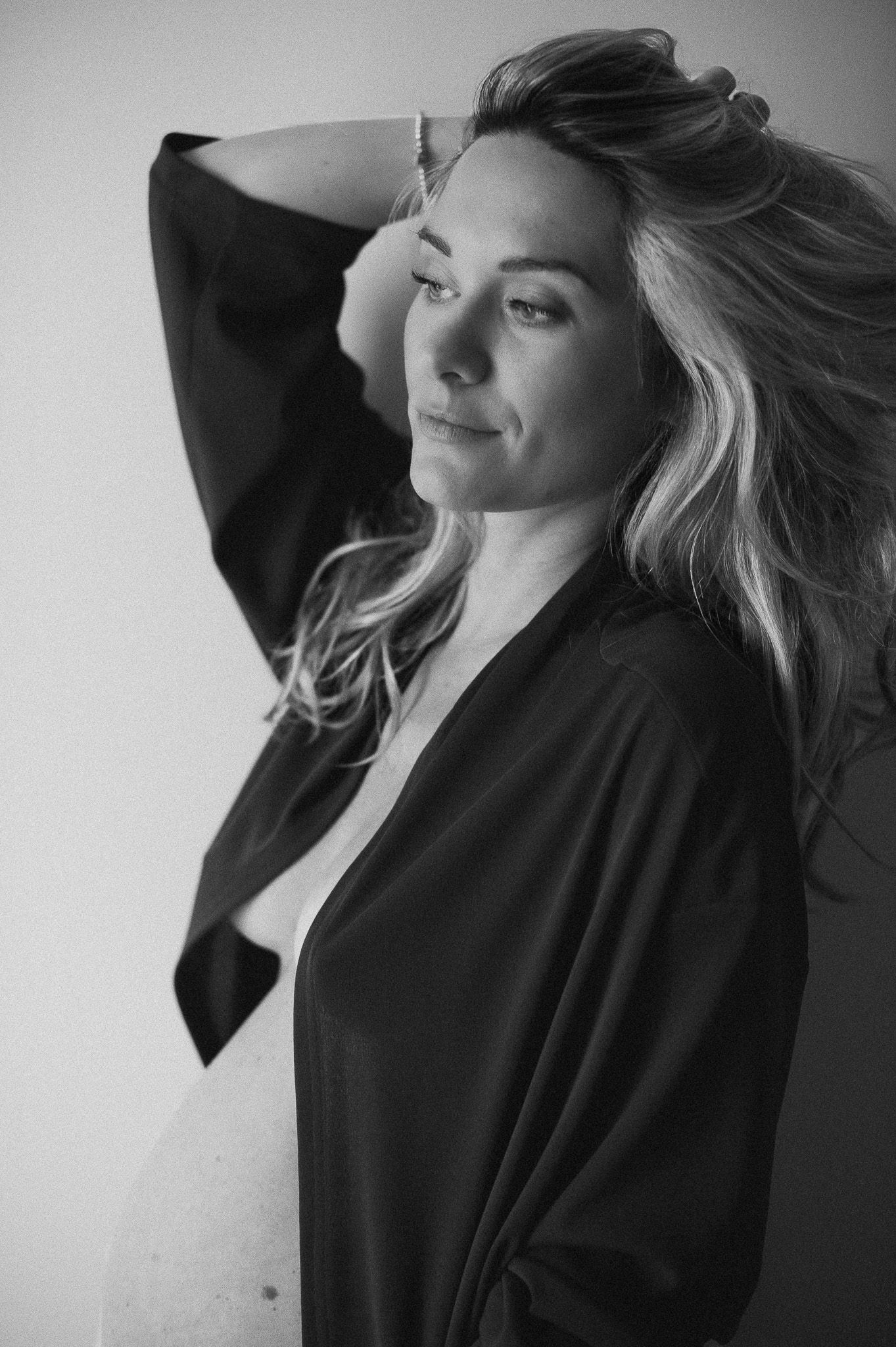 Black and white portrait of a woman with flowing blonde hair in a dark robe, posing gracefully with hand behind head.