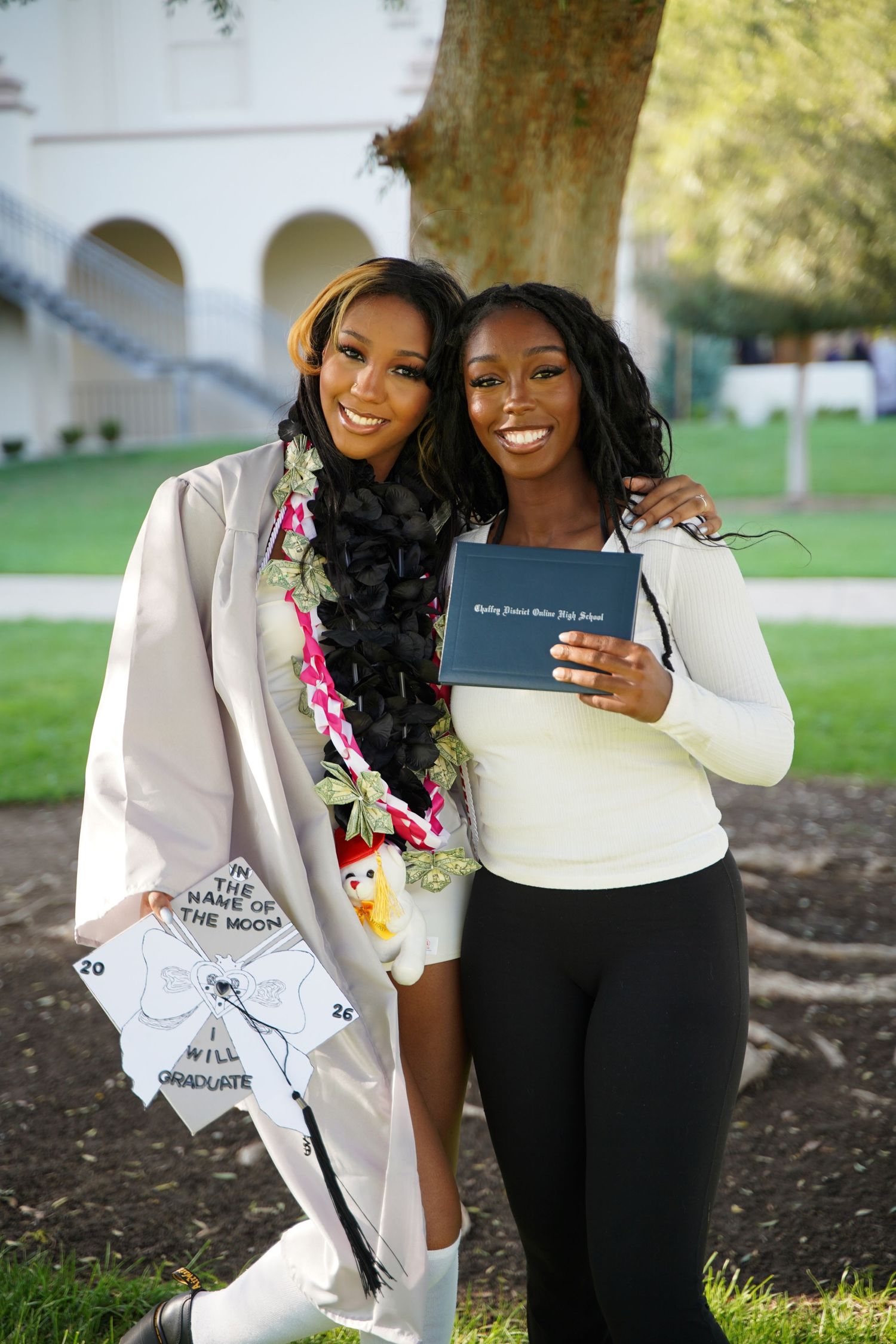 Two young Black women celebrate graduation, one in cap and gown holding diploma at outdoor ceremony.