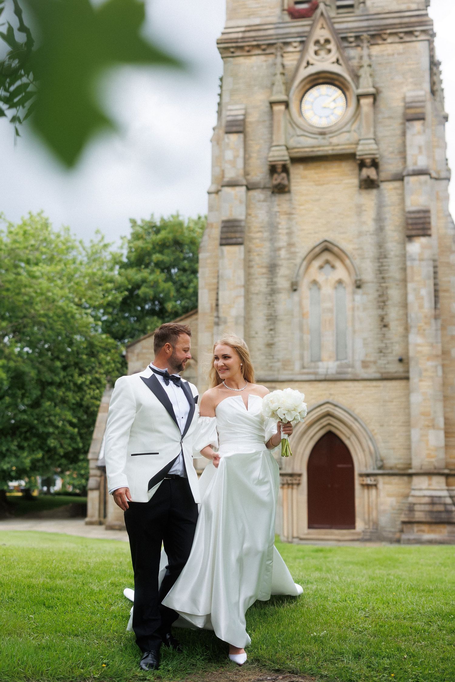 Bride and groom pose outside a Gothic stone church, she in a white gown holding bouquet, he in a white tuxedo jacket.