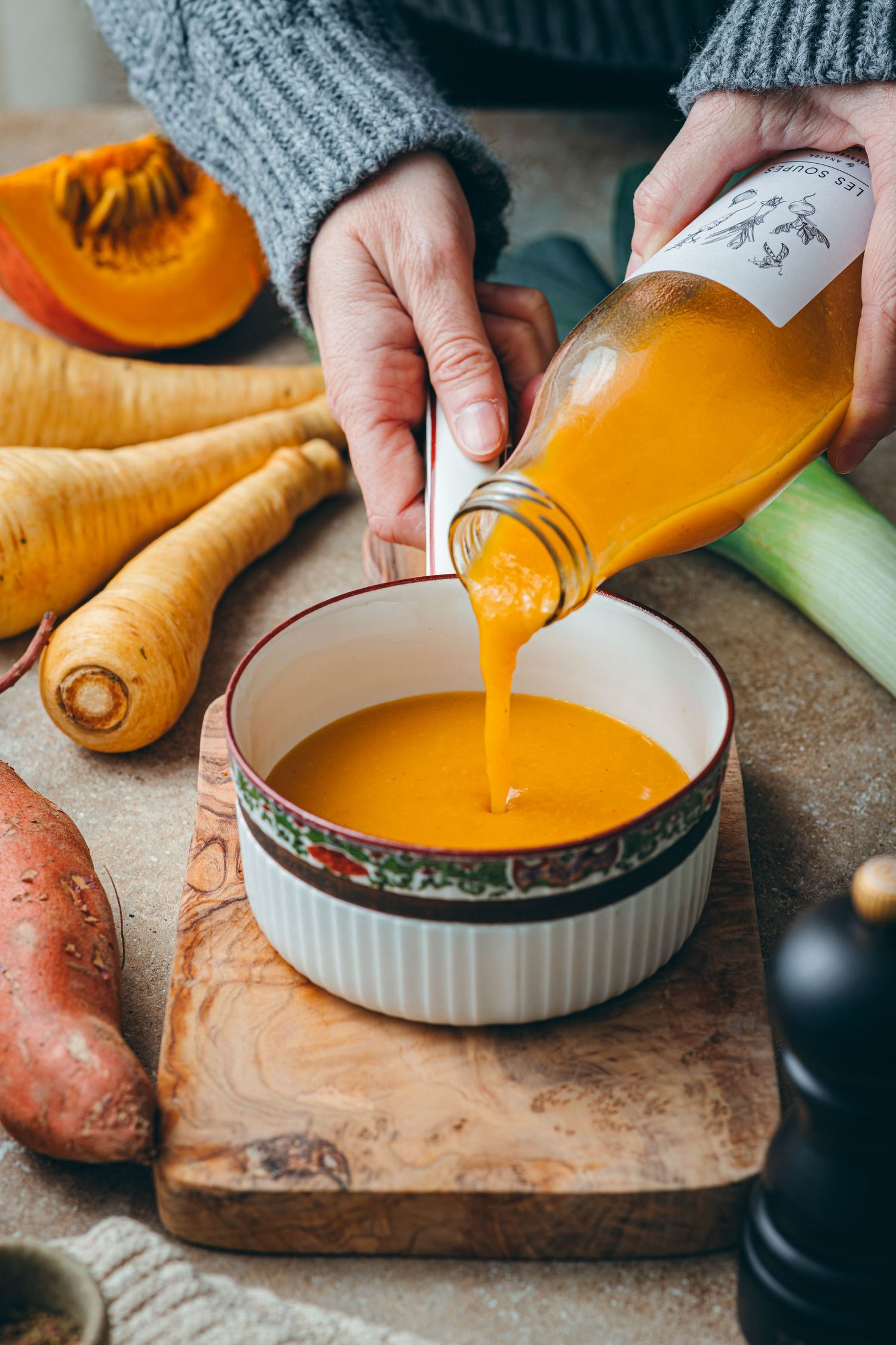 Person pouring orange vegetable soup from bottle into ceramic bowl, surrounded by fresh parsnips, pumpkin, and leeks.
