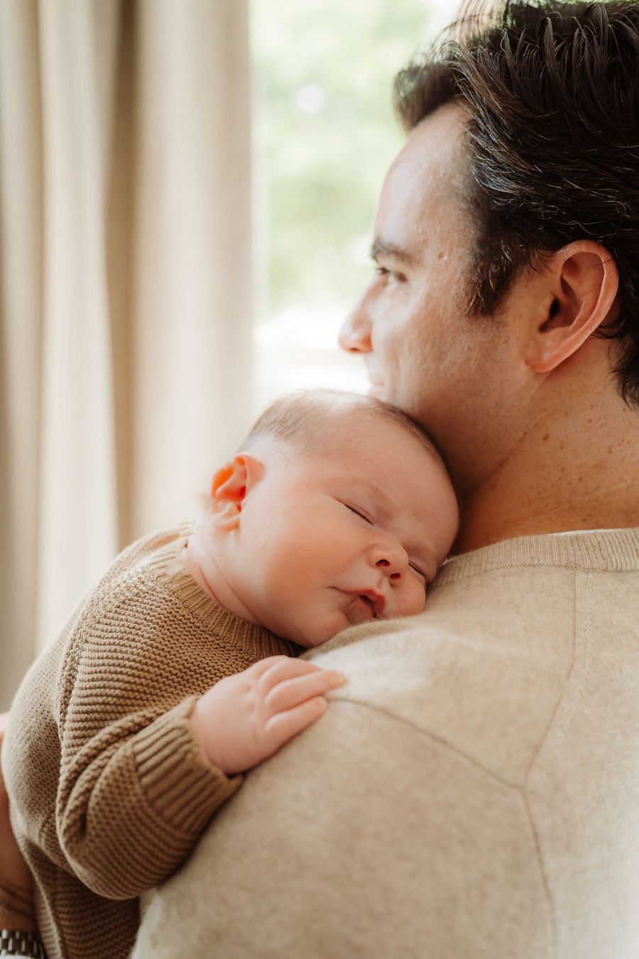 A father tenderly holds his sleeping newborn baby against his shoulder near a bright window.