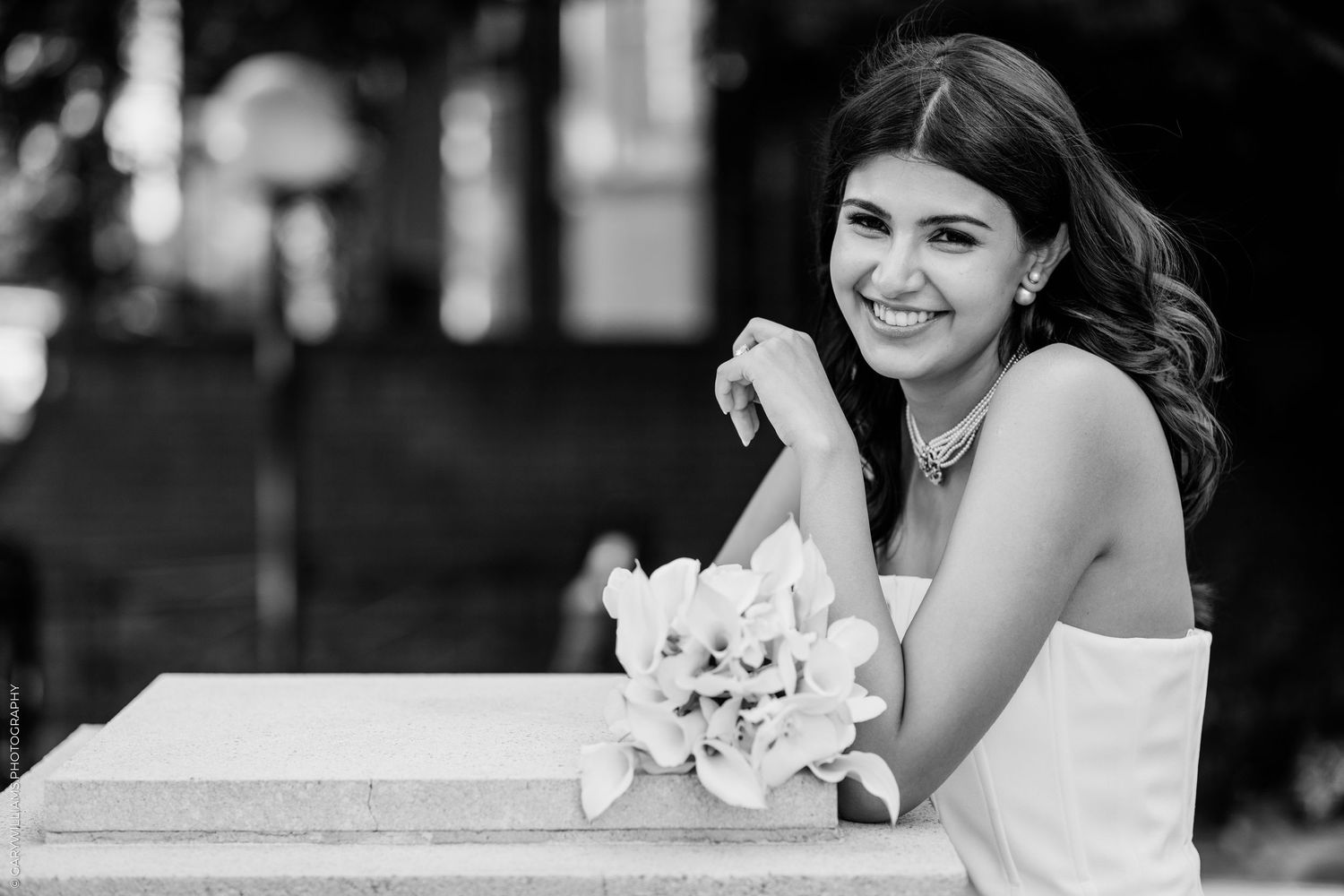 Smiling young woman in white dress holding flowers, leaning on stone ledge outdoors, black and white portrait photo.
