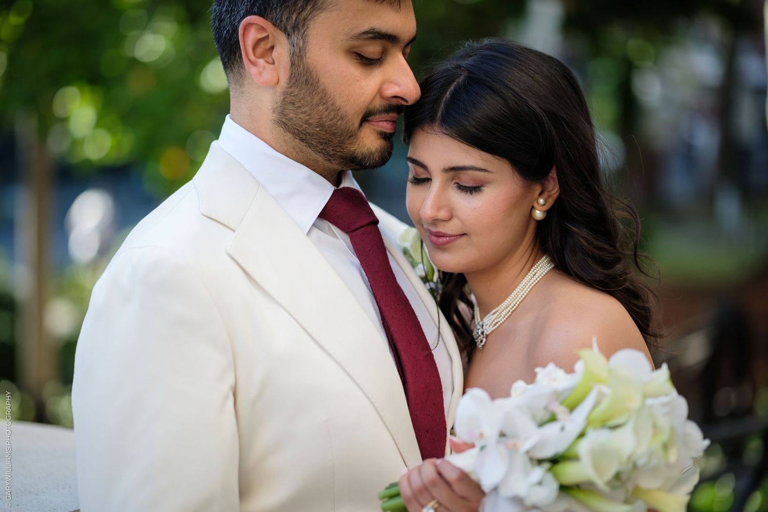 Elegant couple in wedding attire, groom in white suit with red tie, bride holding white floral bouquet, sharing a tender moment.