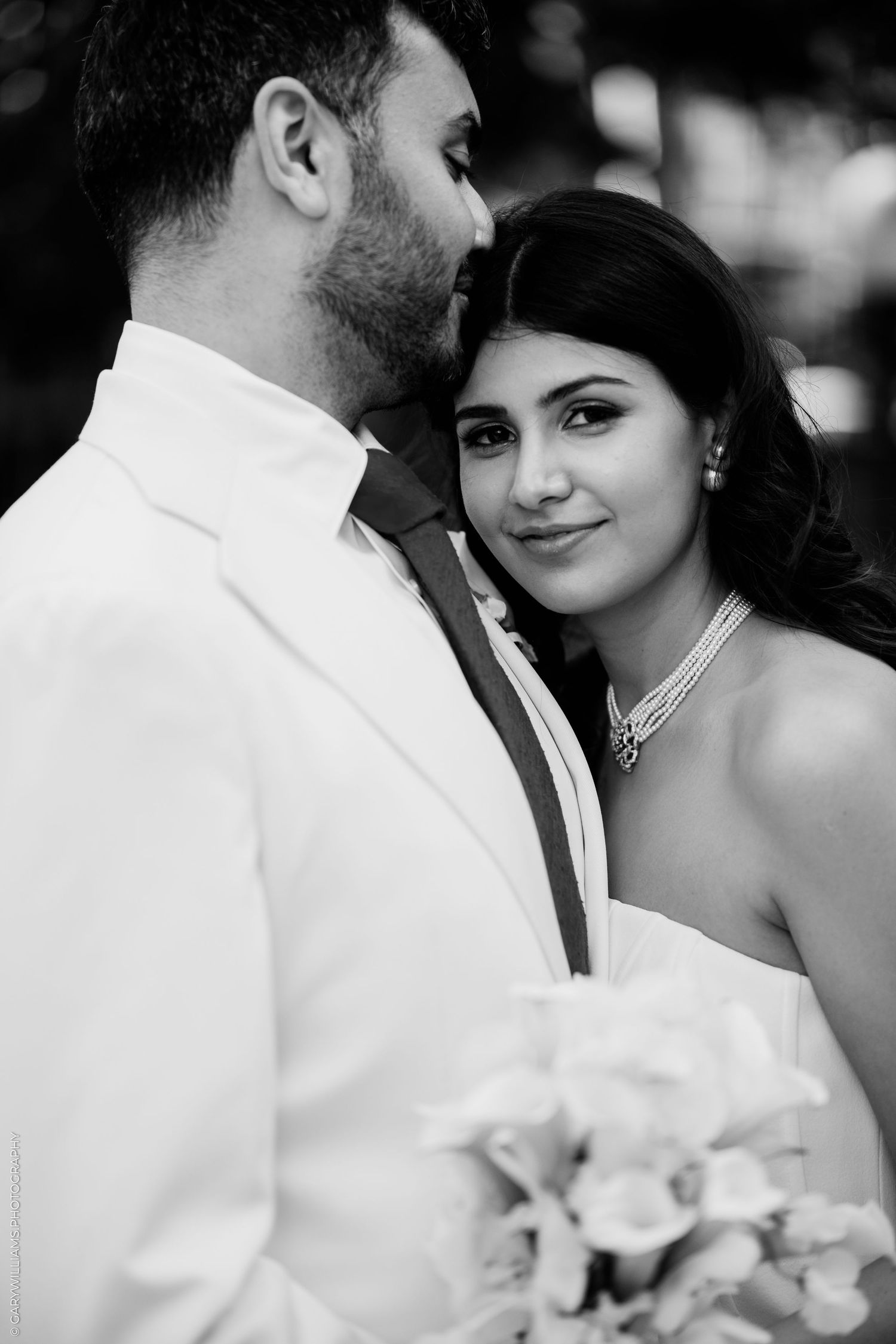 Black and white wedding portrait of bride and groom in intimate pose, bride wearing necklace holding bouquet.