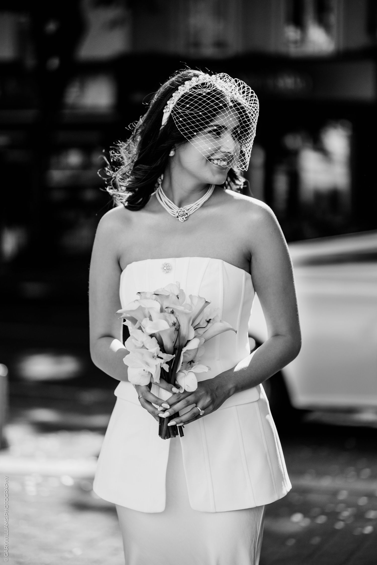 Black and white photo of a smiling bride in strapless gown, birdcage veil, holding floral bouquet outdoors.
