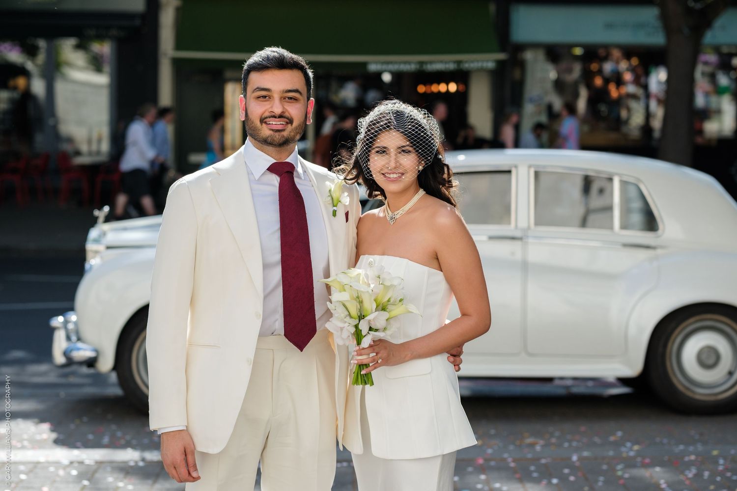 Bride and groom posing in front of a classic white Rolls-Royce on their wedding day, bride holding white flowers.