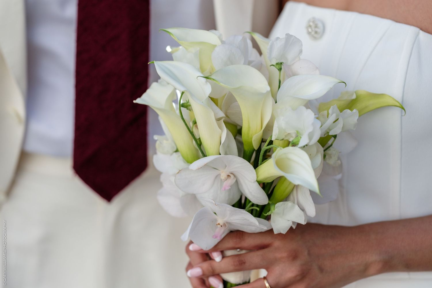 Bride holding elegant white calla lily wedding bouquet, with groom in white suit and red tie visible in background.