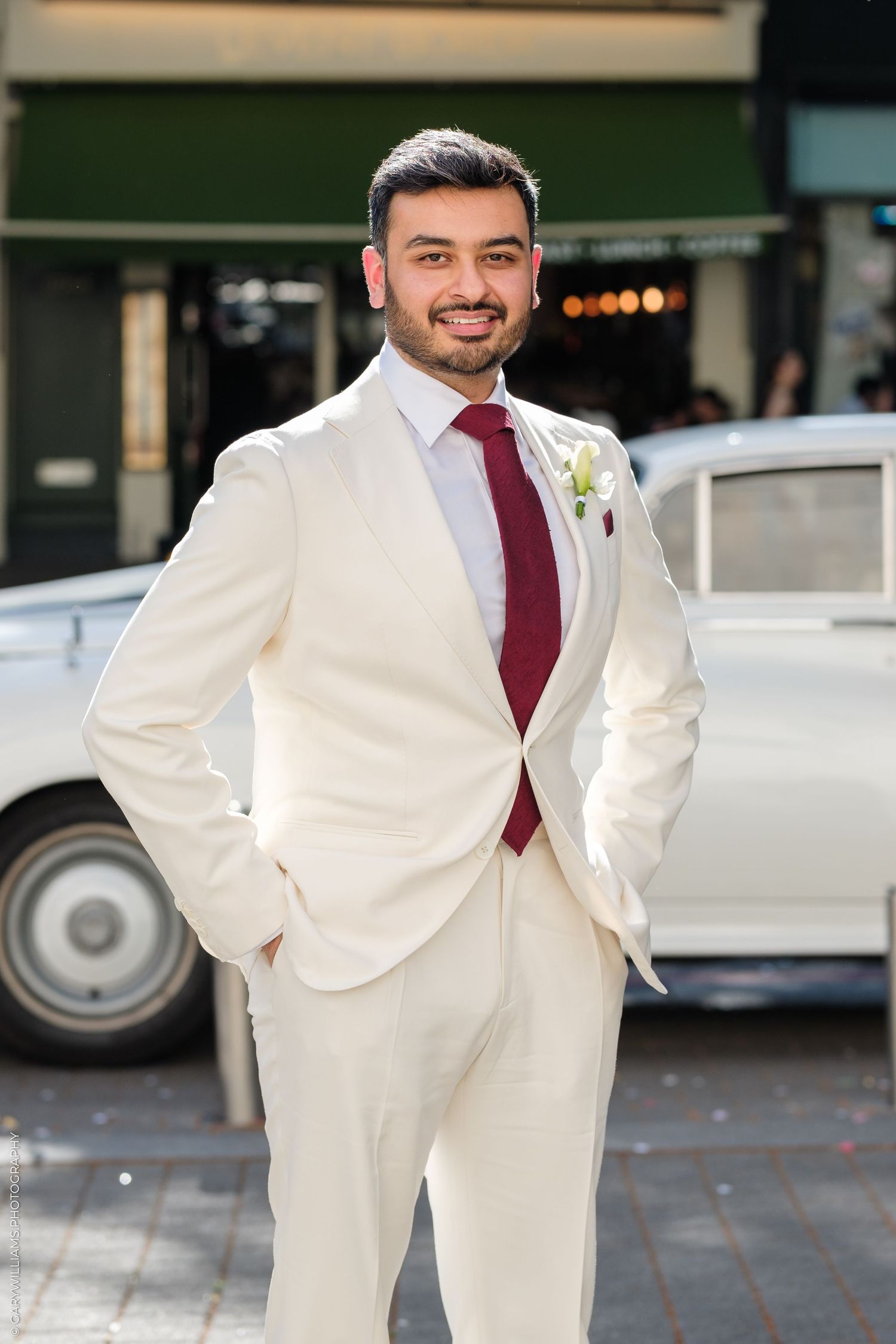 Groom in cream suit with burgundy tie posing confidently outdoors beside a vintage white car.