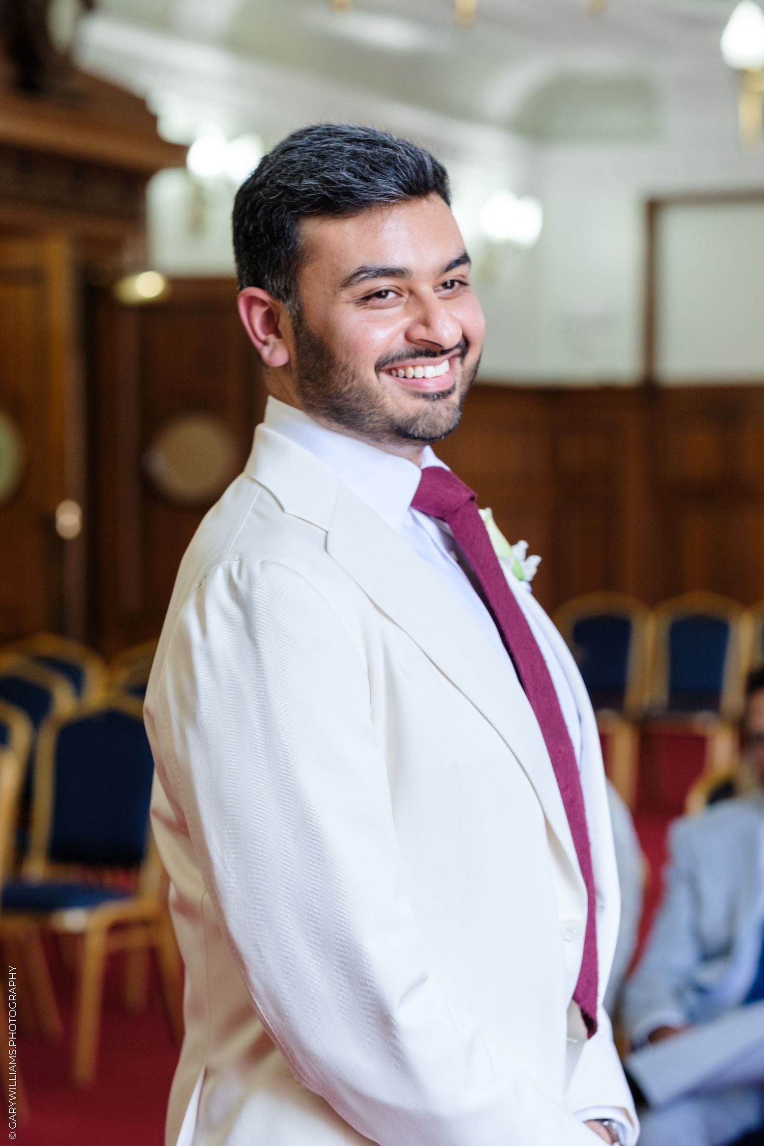 Smiling groom in cream wedding suit with burgundy tie standing in elegant ceremony venue interior.