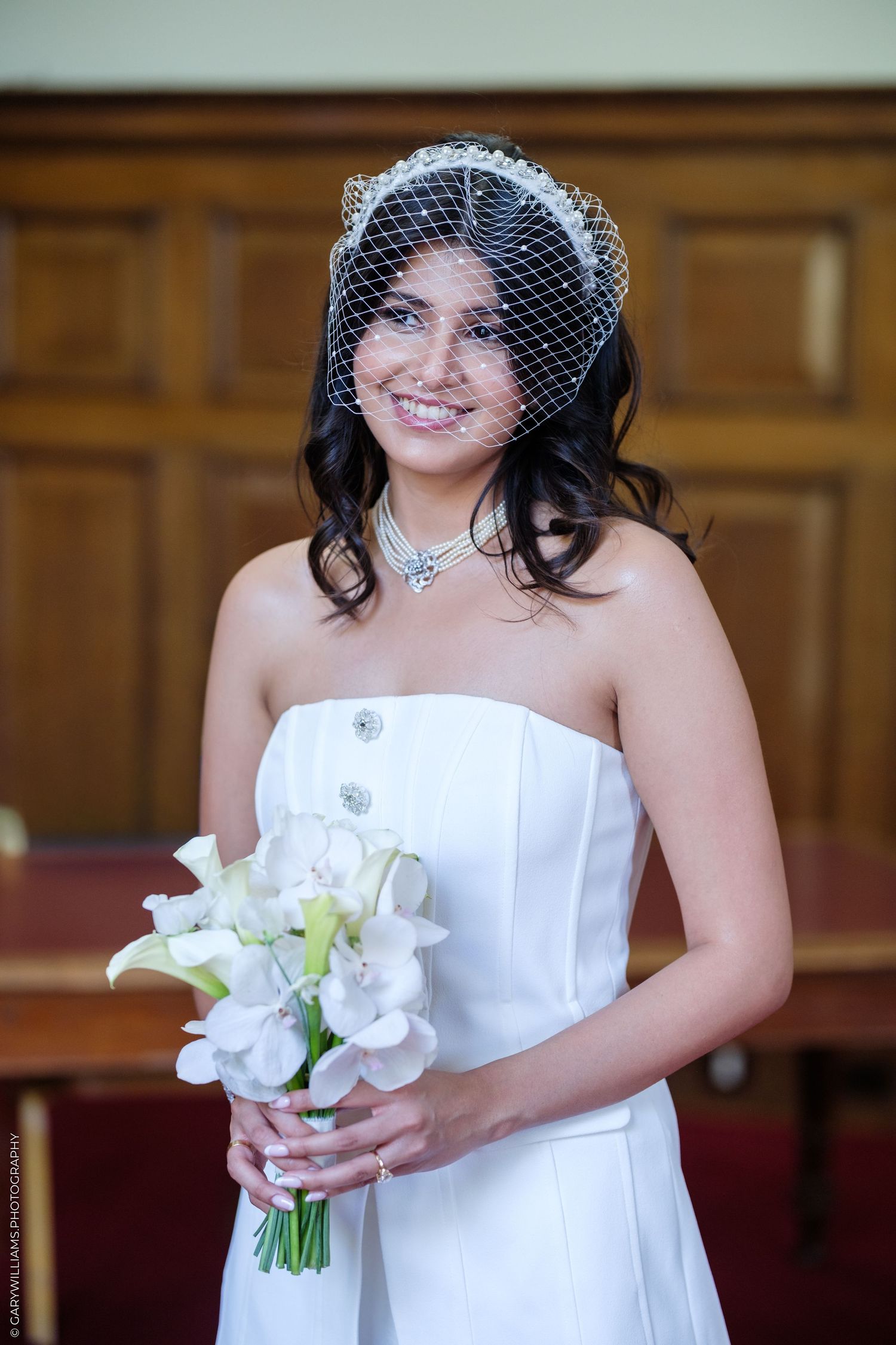 Smiling bride in strapless white gown and birdcage veil holds white floral bouquet indoors.