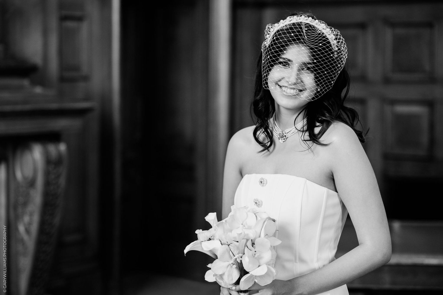Black and white portrait of bride in strapless gown wearing birdcage veil holding floral bouquet.