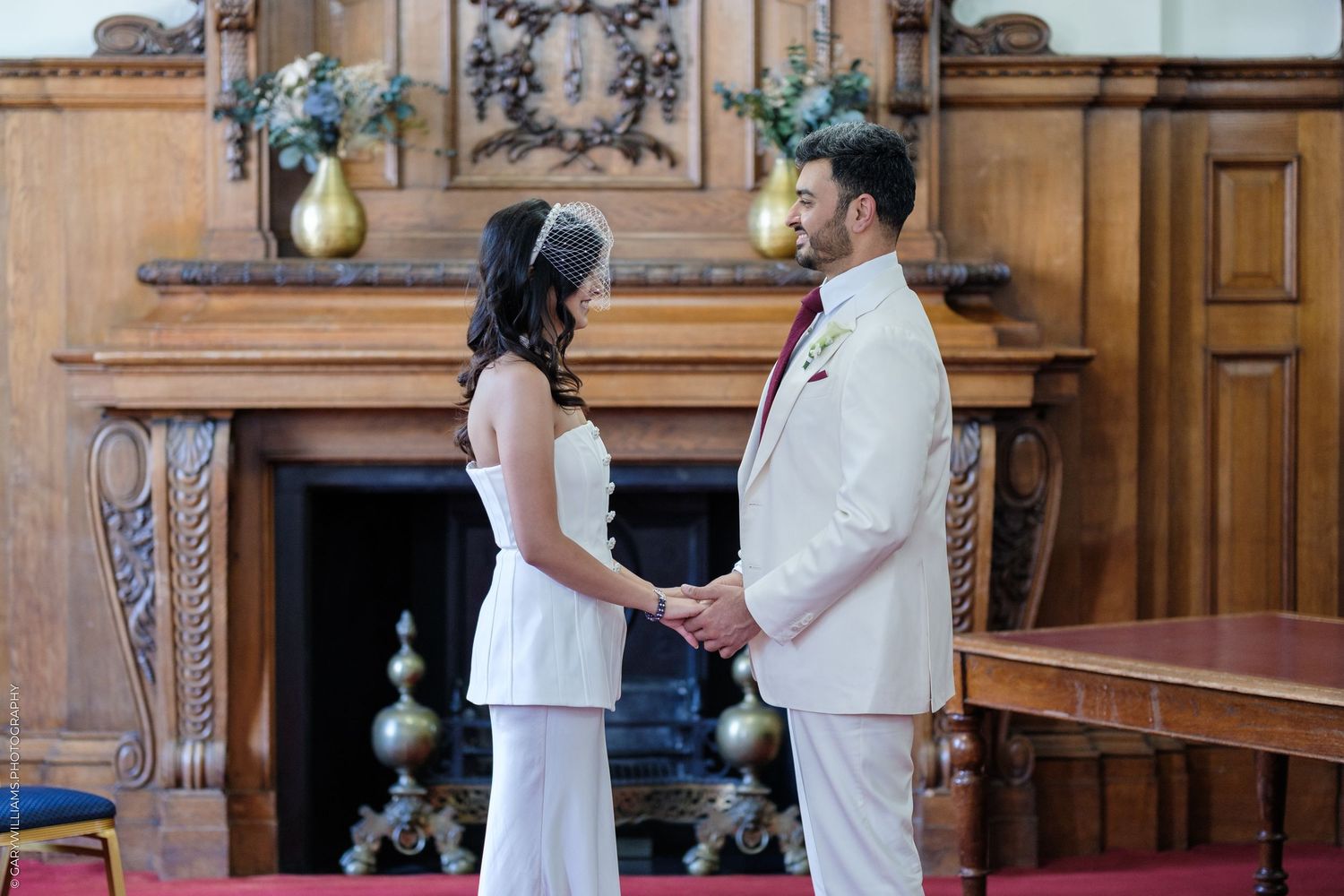 Bride and groom holding hands at wedding ceremony in elegant wood-paneled room with ornate fireplace.