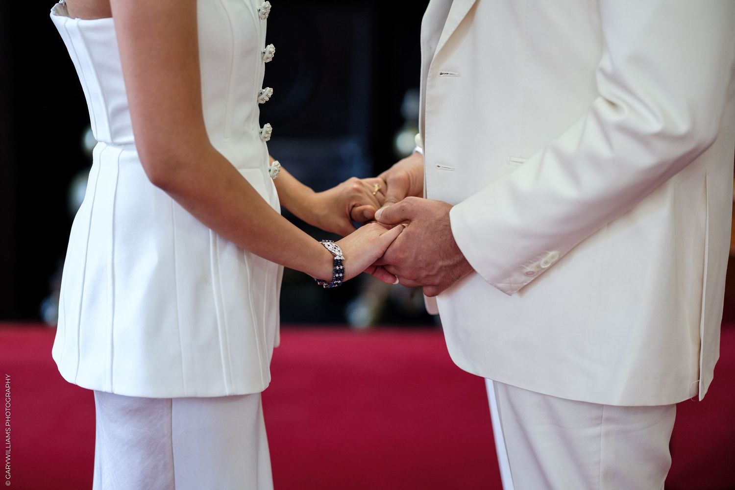 Couple in white attire holding hands during a wedding ceremony on a red carpet.