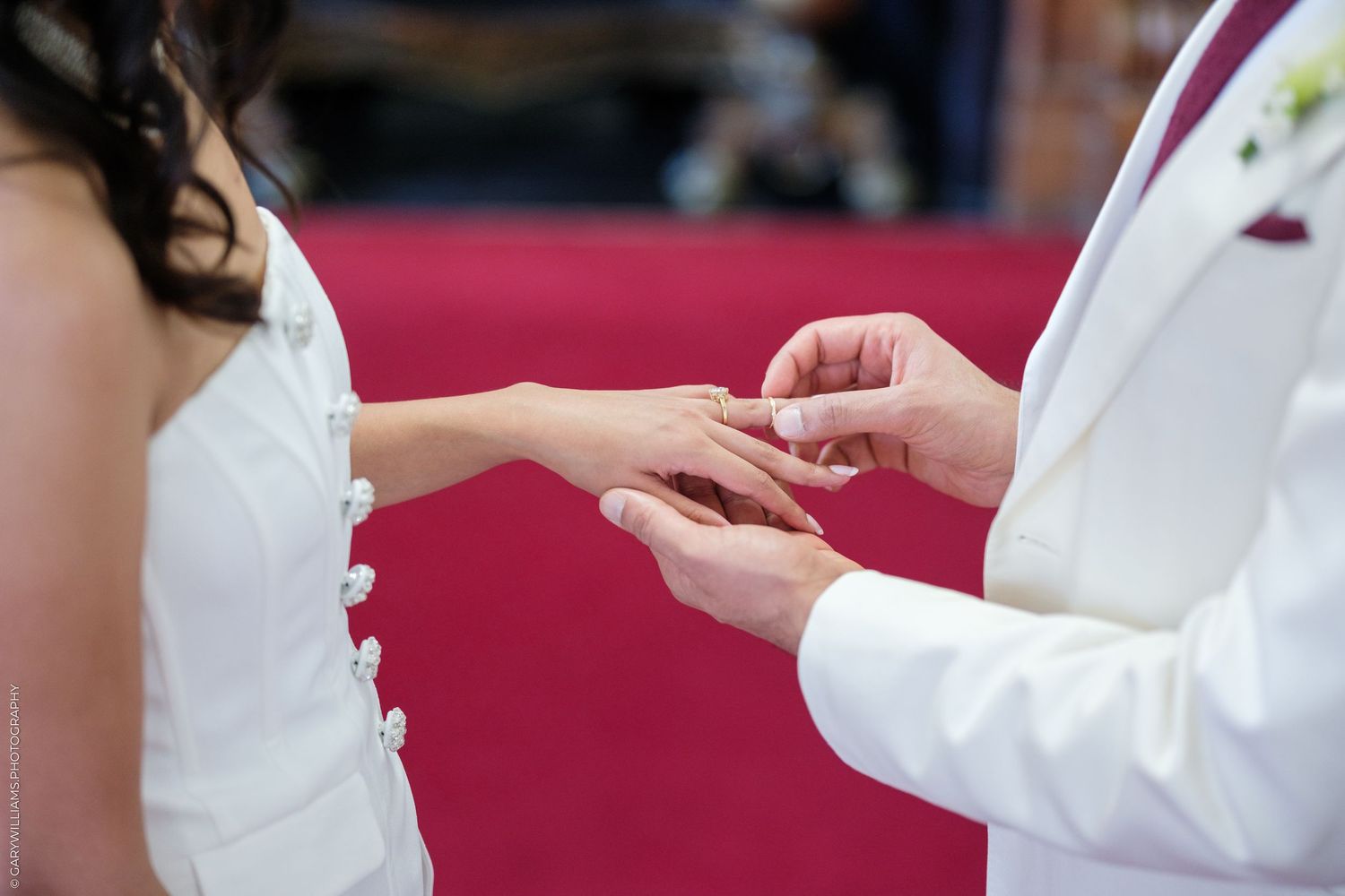 Groom in white suit placing wedding ring on bride's finger during marriage ceremony.