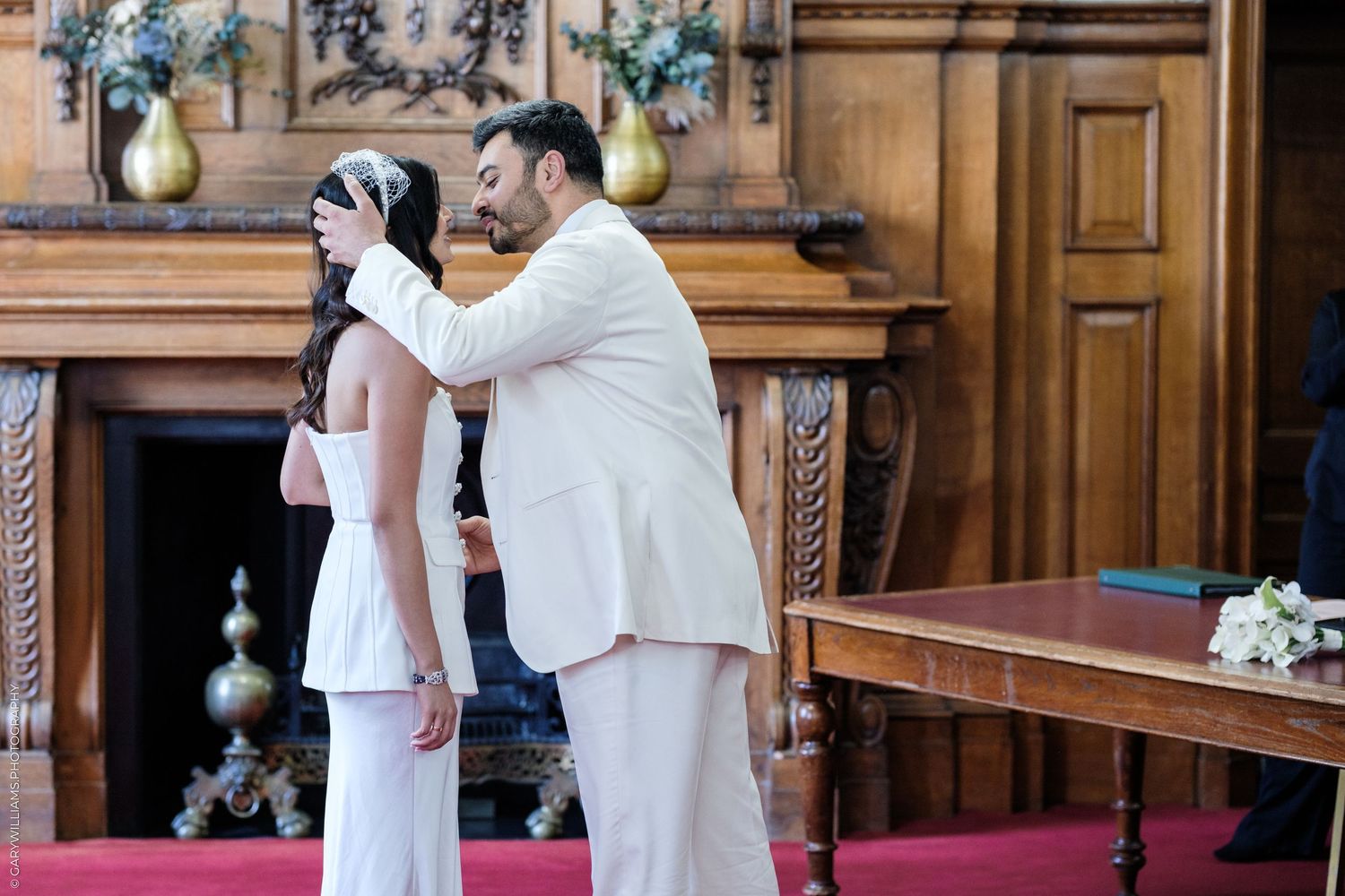 Groom in white suit lifting bride's veil during intimate wedding ceremony in elegant wood-paneled room.