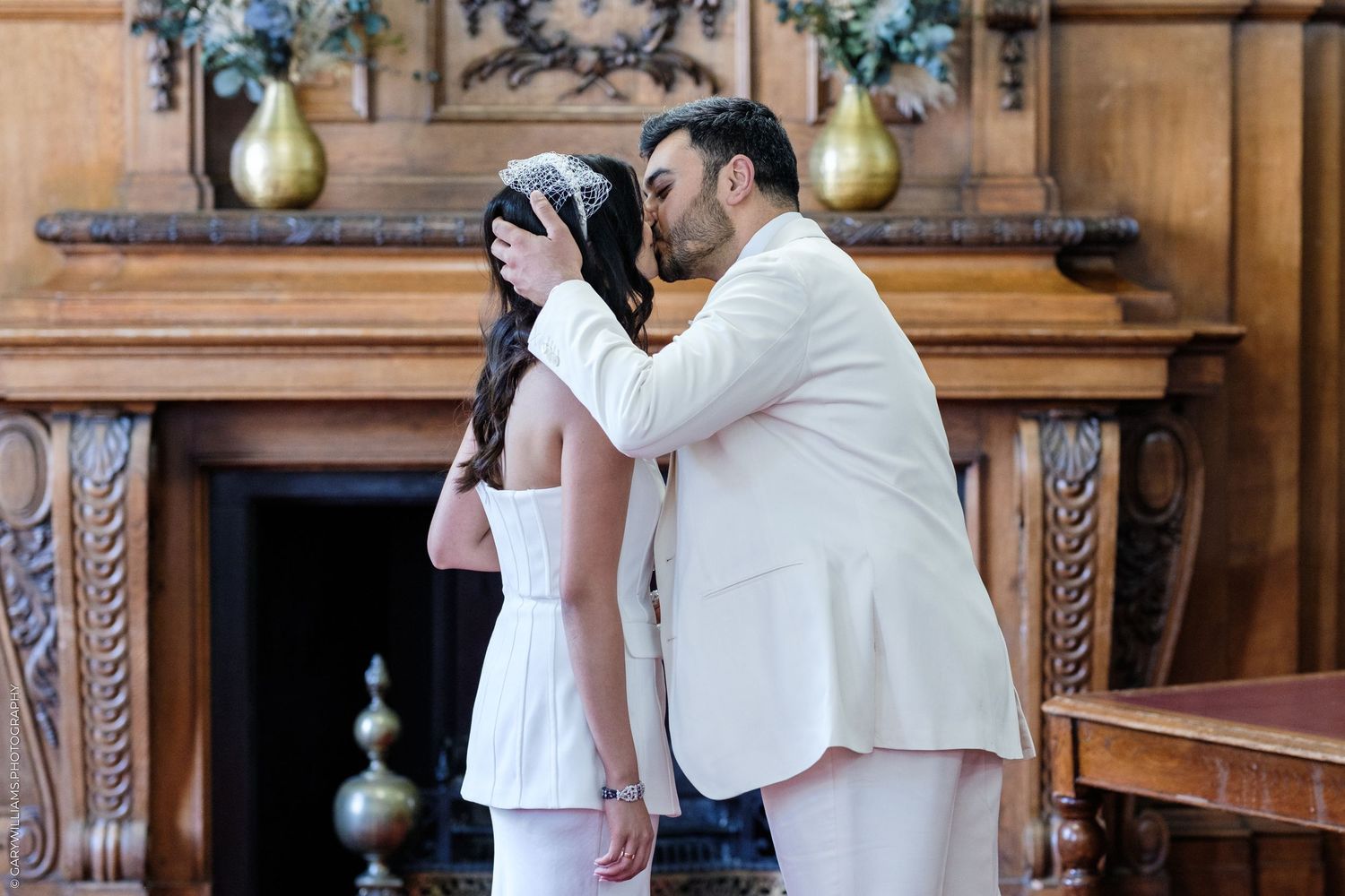 Groom in white suit kissing bride in white dress near ornate wooden fireplace during wedding ceremony.