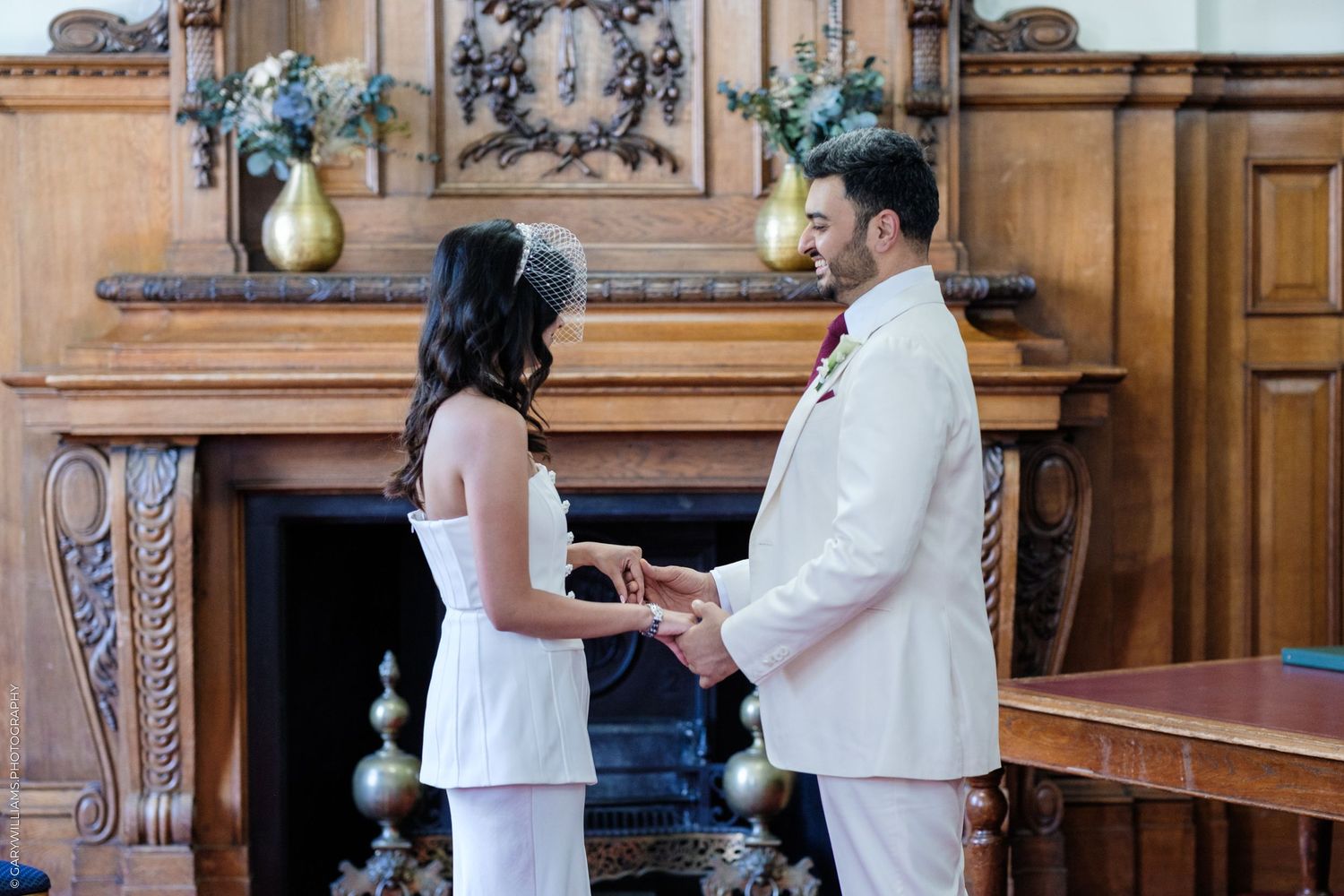 Bride and groom exchange rings at elegant wood-paneled fireplace ceremony in white wedding attire.