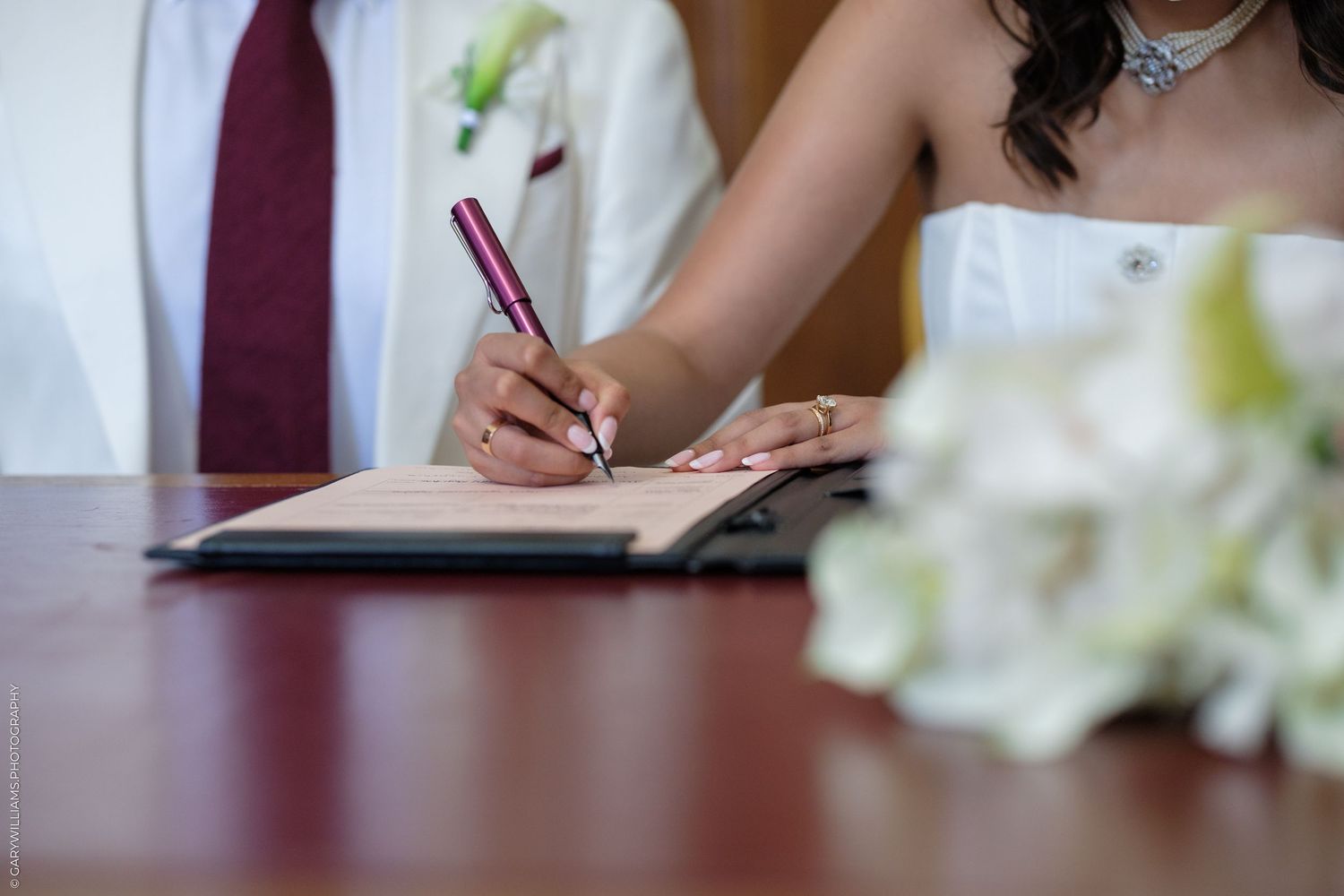 Bride signing wedding registry document with a pink pen, groom beside her, bouquet in foreground.