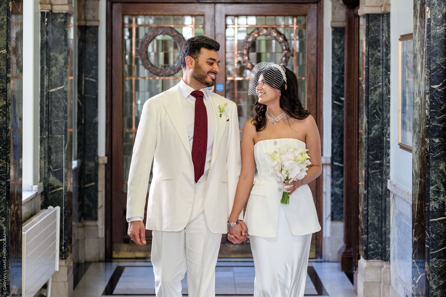 Bride and groom holding hands walking in elegant marble hallway, bride in strapless white gown holding bouquet, groom in white suit.