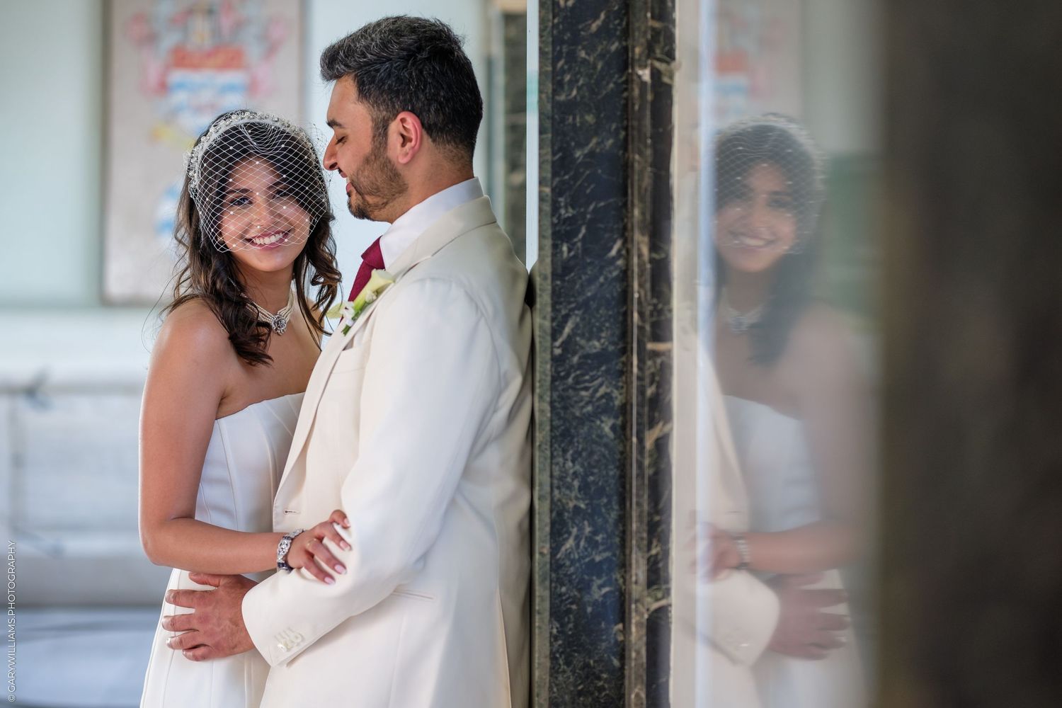 A bride and groom share a tender embrace near a marble column, their reflection visible in a mirror beside them.