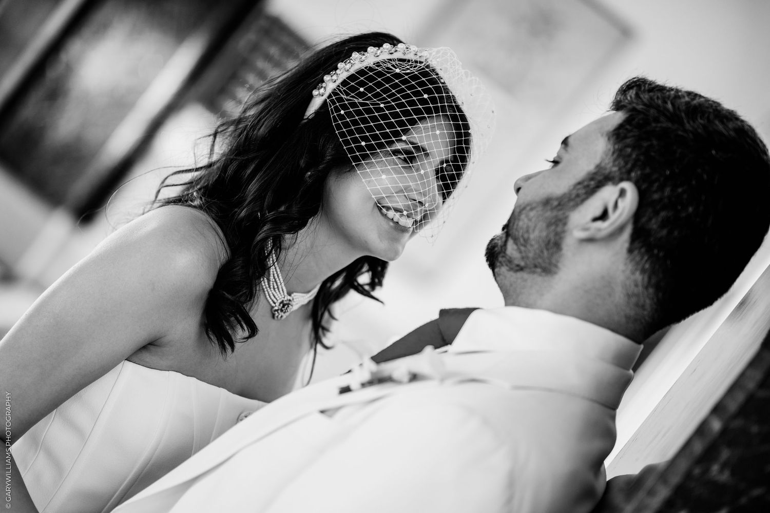 Black and white photo of a bride in a birdcage veil smiling at her groom in an intimate wedding moment.