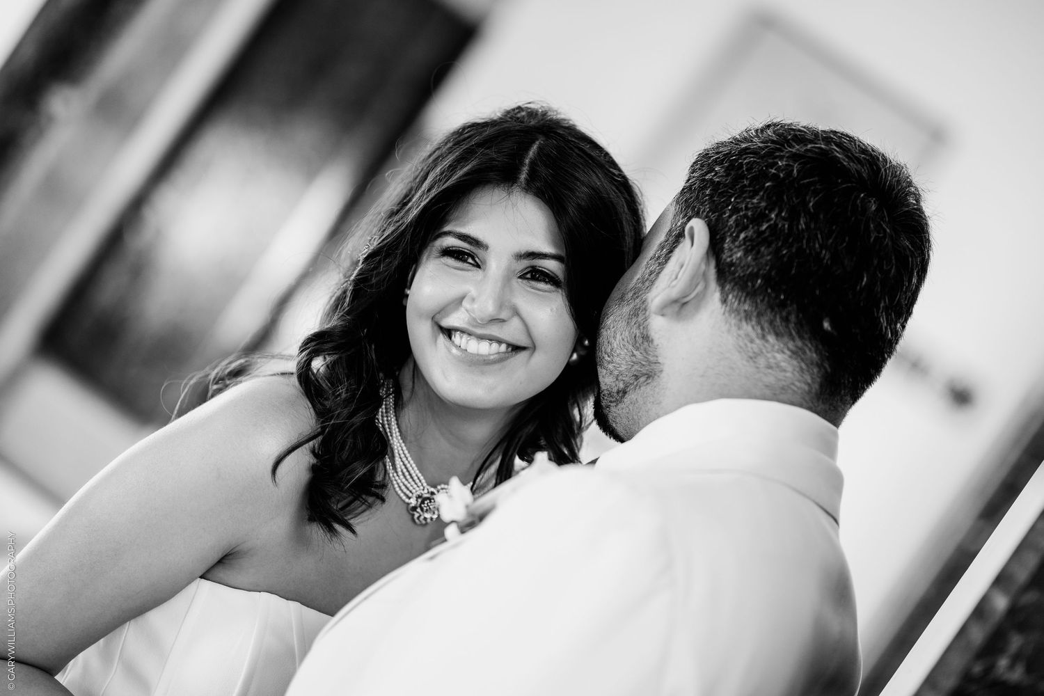 Black and white wedding photo of a smiling bride wearing a statement necklace as groom kisses her cheek.