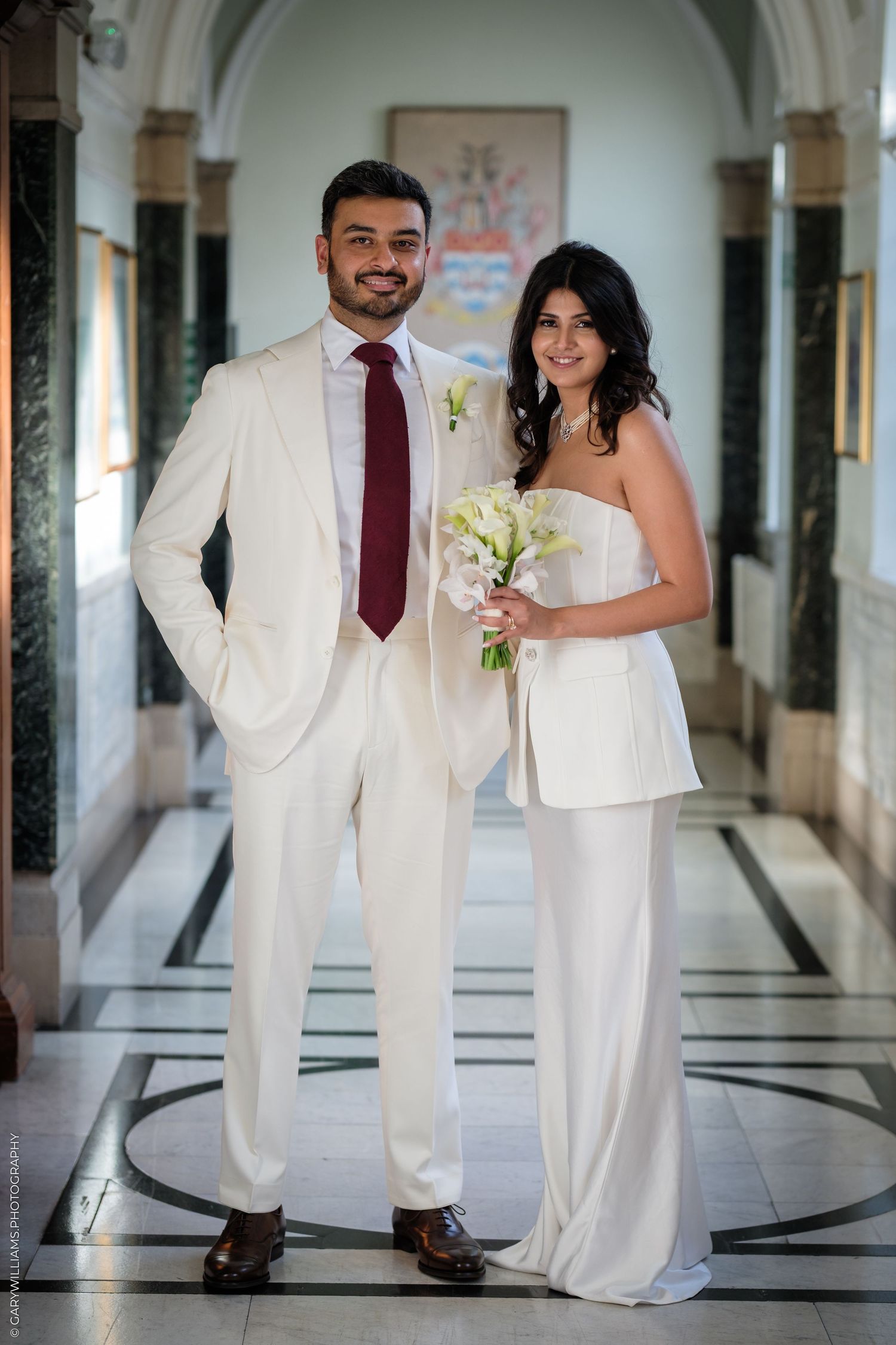 Bride in white jumpsuit holding bouquet stands with groom in cream suit and burgundy tie in elegant marble hallway.