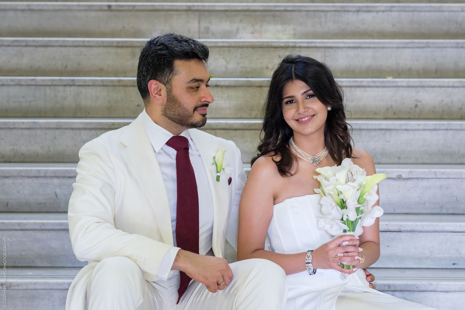 A couple in wedding attire sits on stone steps, groom in white suit with red tie, bride holding white calla lily bouquet.