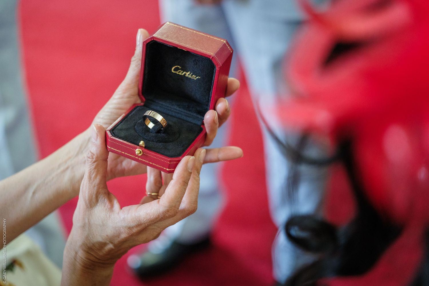 Hands presenting an open red Cartier ring box with a gold ring inside at a proposal or special occasion.