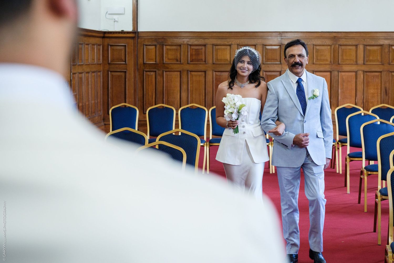Bride in white dress walks down aisle with suited man in elegant wood-paneled ceremony hall with blue chairs.