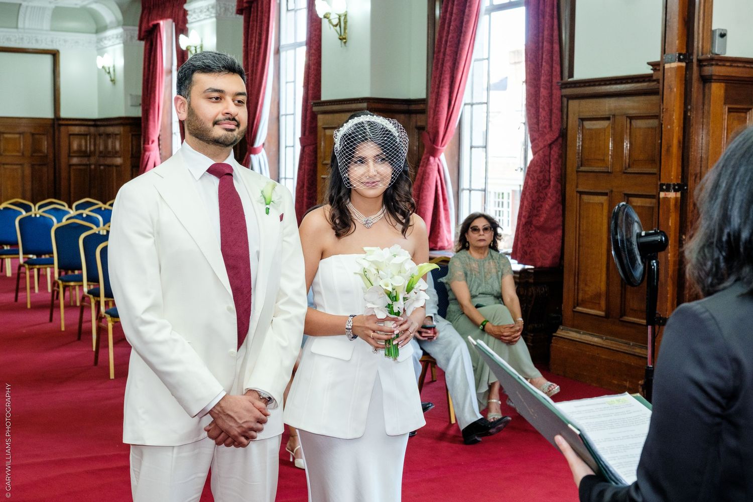 Bride and groom stand together at civil wedding ceremony in elegant red-curtained room, bride holding white bouquet.
