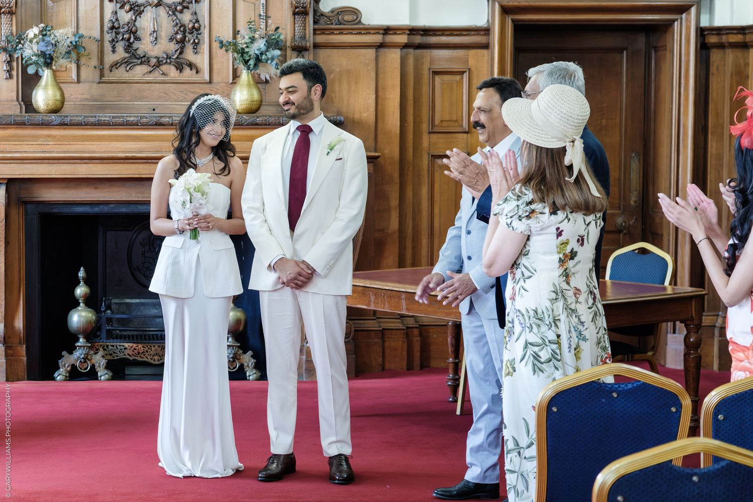 Couple in white wedding attire smiling at civil ceremony in elegant wood-paneled room with guests applauding.
