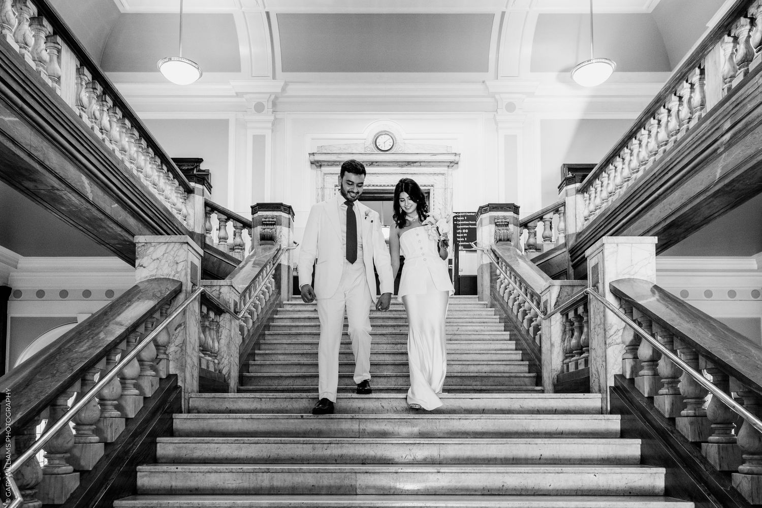 Black and white photo of a couple holding hands on grand staircase in elegant building.