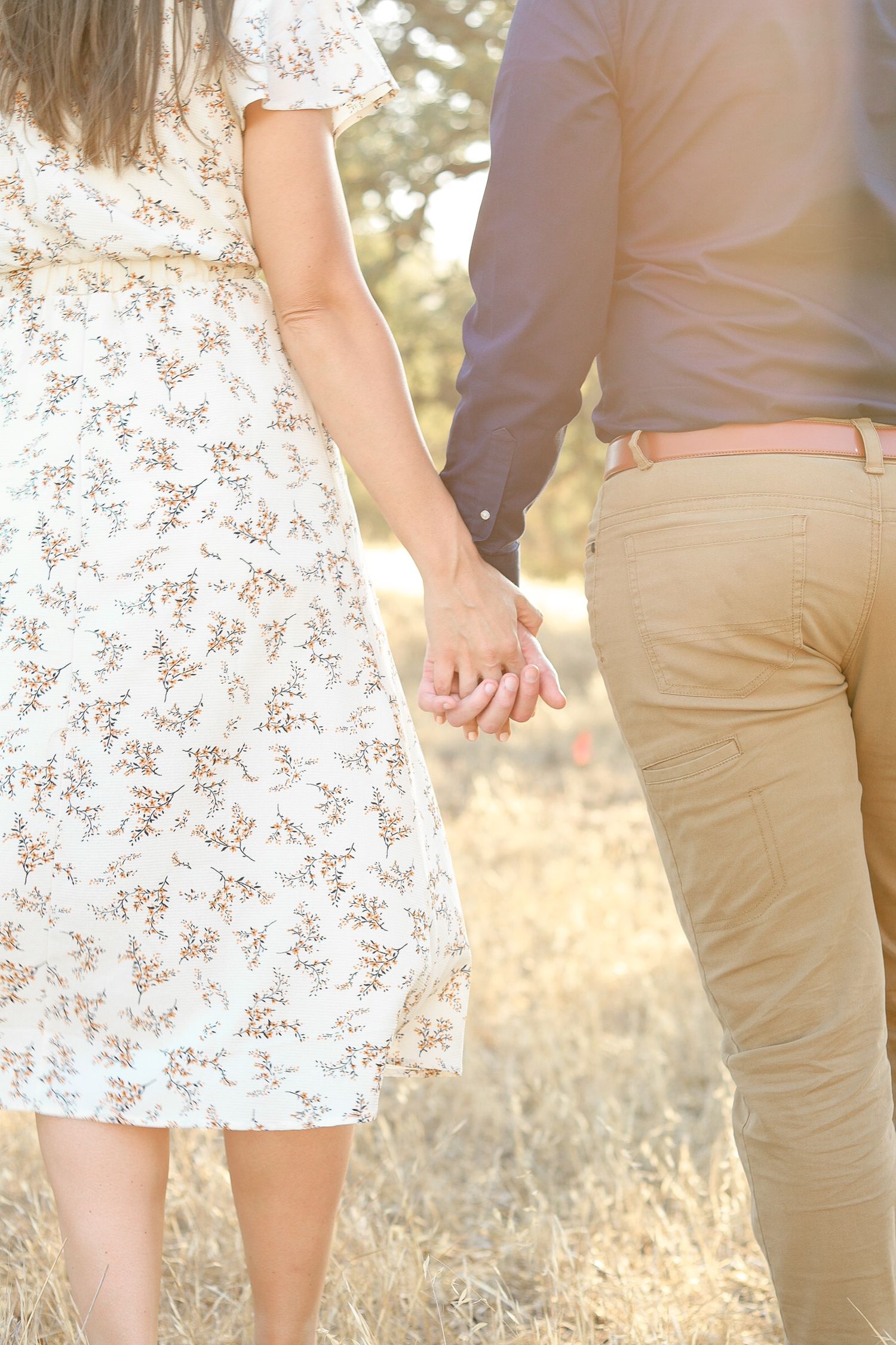 Couple holding hands in a sunny field, woman in floral dress and man in khaki pants walking together outdoors.