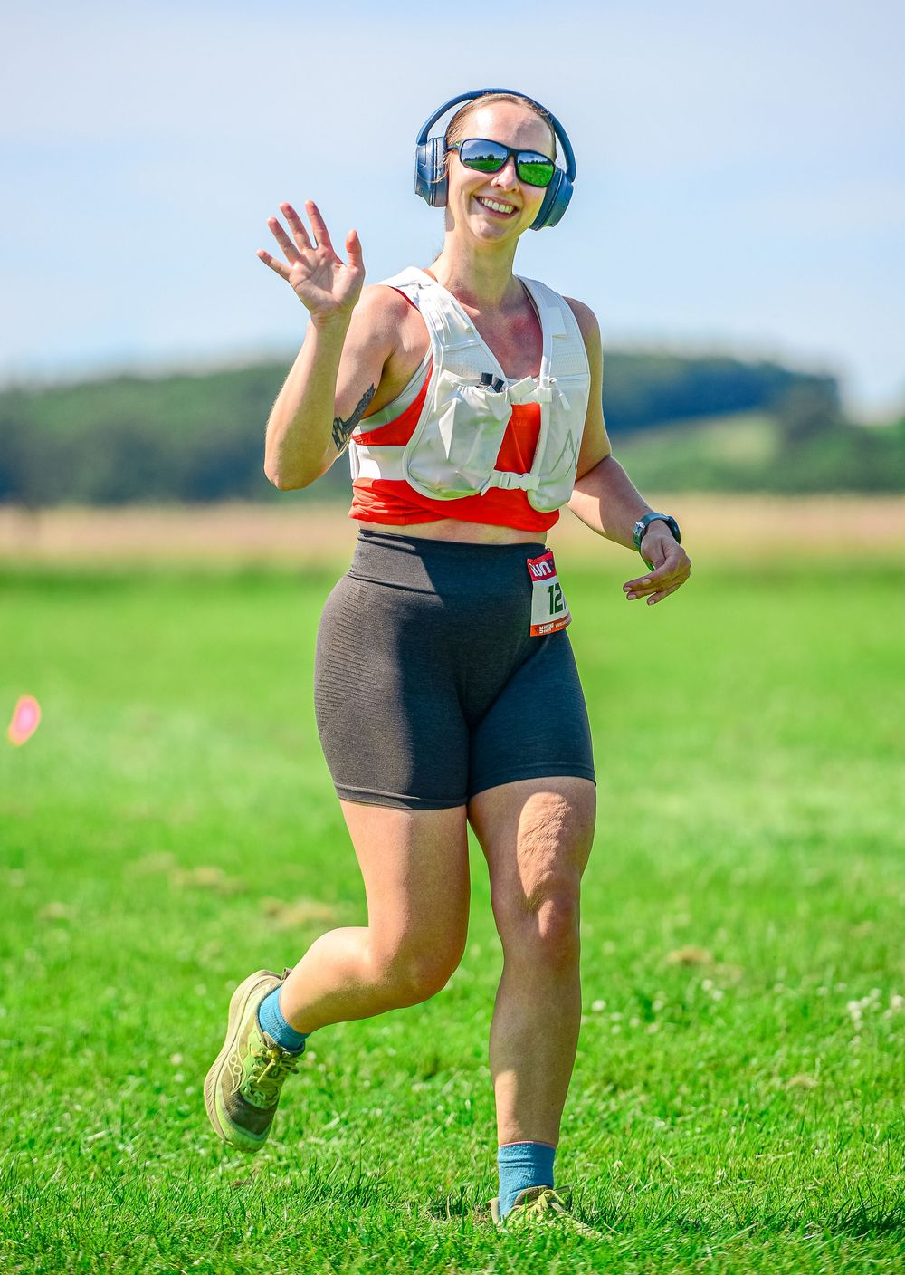 Smiling woman runner wearing headphones and hydration vest waves during outdoor race on sunny day.