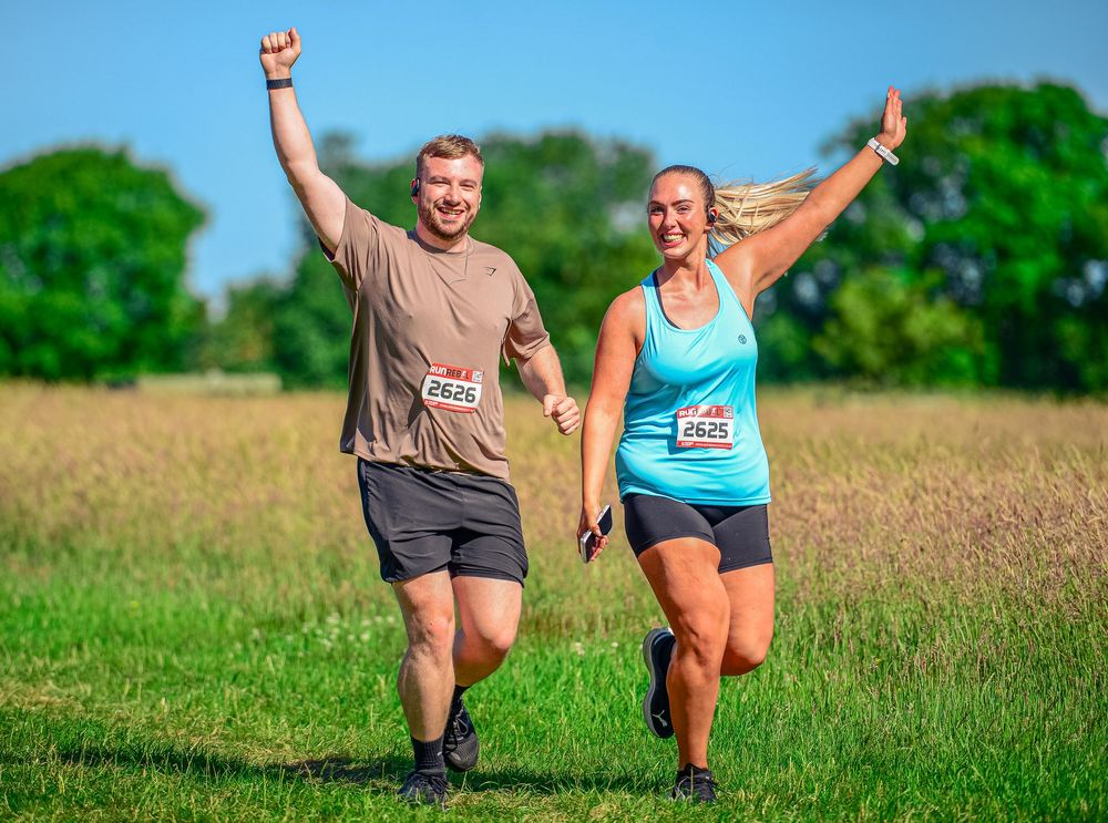 Cheerful man and woman with race bibs run together through sunny green field at outdoor event.