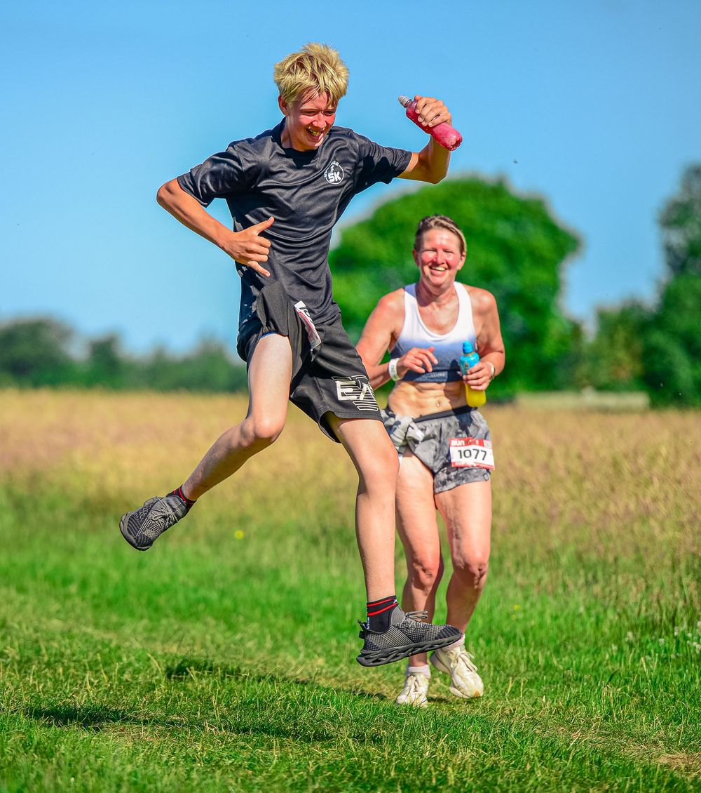 Two energetic runners laugh and leap through grassy field during outdoor race on a sunny day.