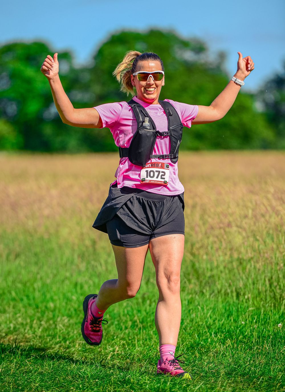 Happy woman runner in pink shirt gives thumbs up wearing race bib 1072 in sunny green field.