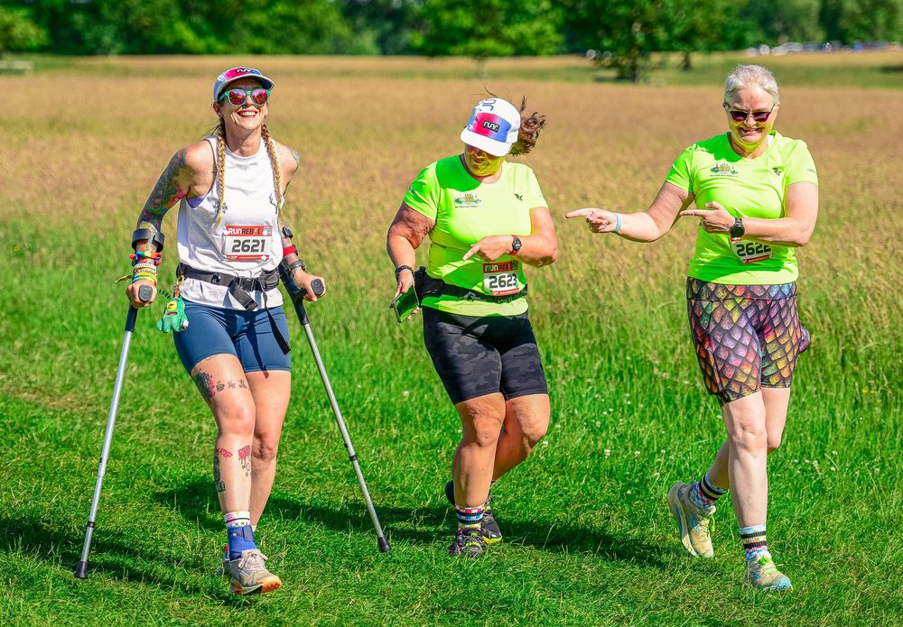 Three women walk together using trekking poles during outdoor charity race event on sunny day.