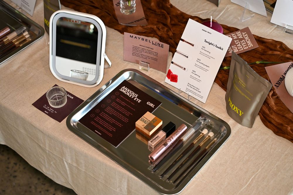 Maybelline makeup display with mirror, cosmetics on silver tray, and branded signage on a beige table.
