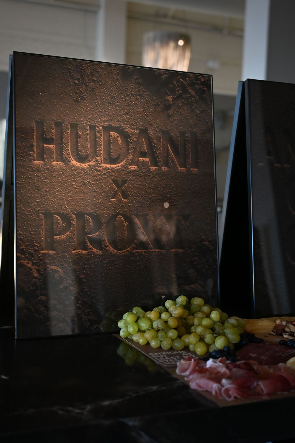 Hudani x Proxy branded stone plaque display with fresh green grapes and charcuterie on a dark table.