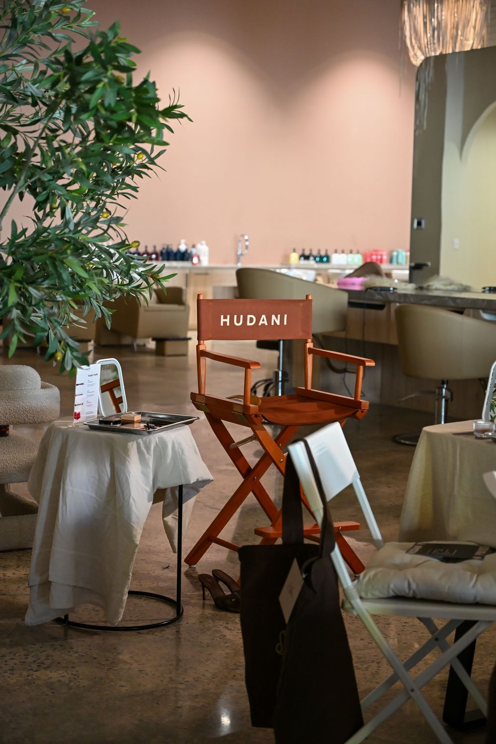 A director's chair labeled HUDANI sits in a modern salon with nail stations, a potted tree, and elegant white draped tables.