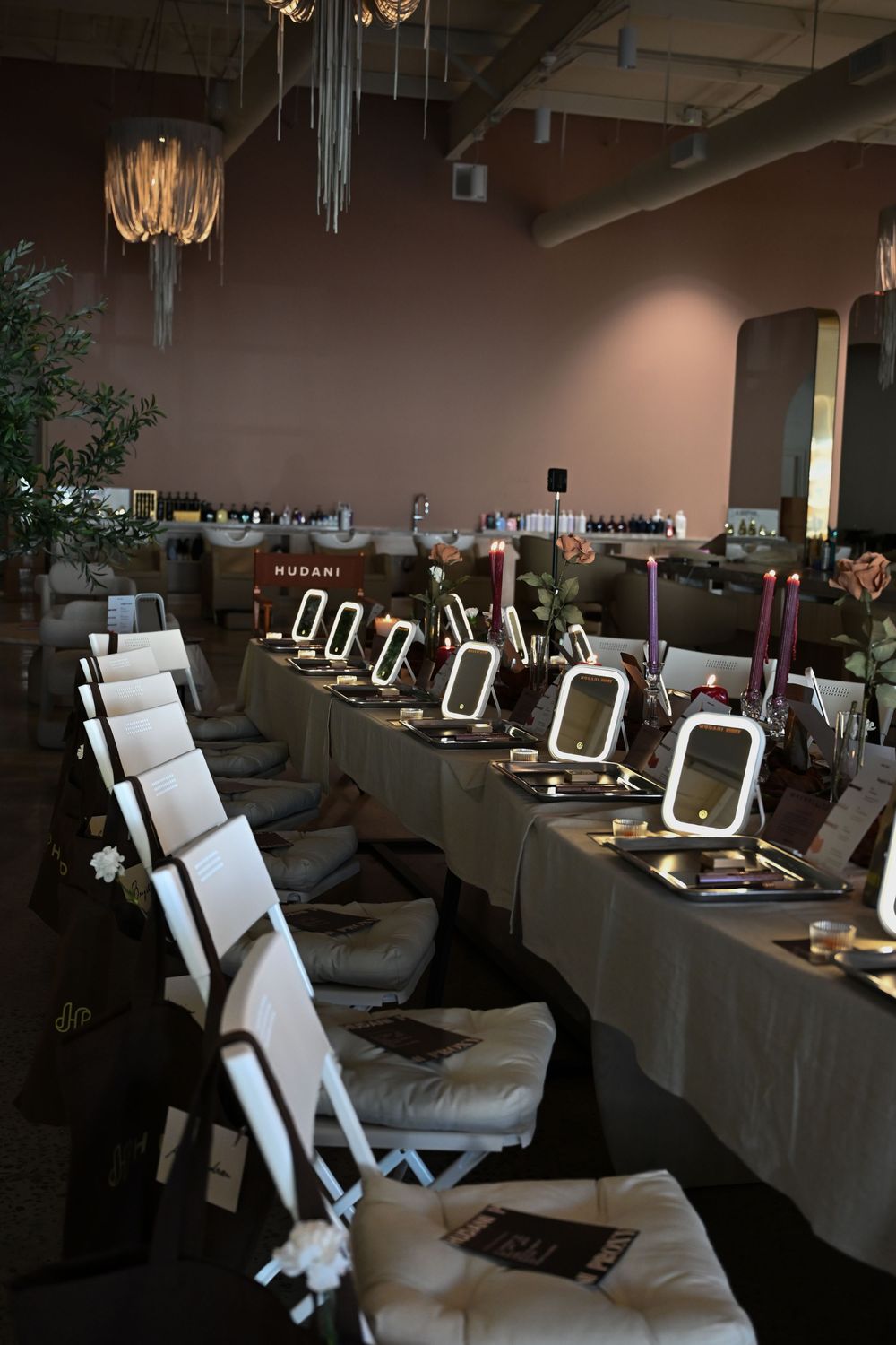 A long makeup station table with lit mirrors and white chairs set up in an elegant salon event space with chandeliers.