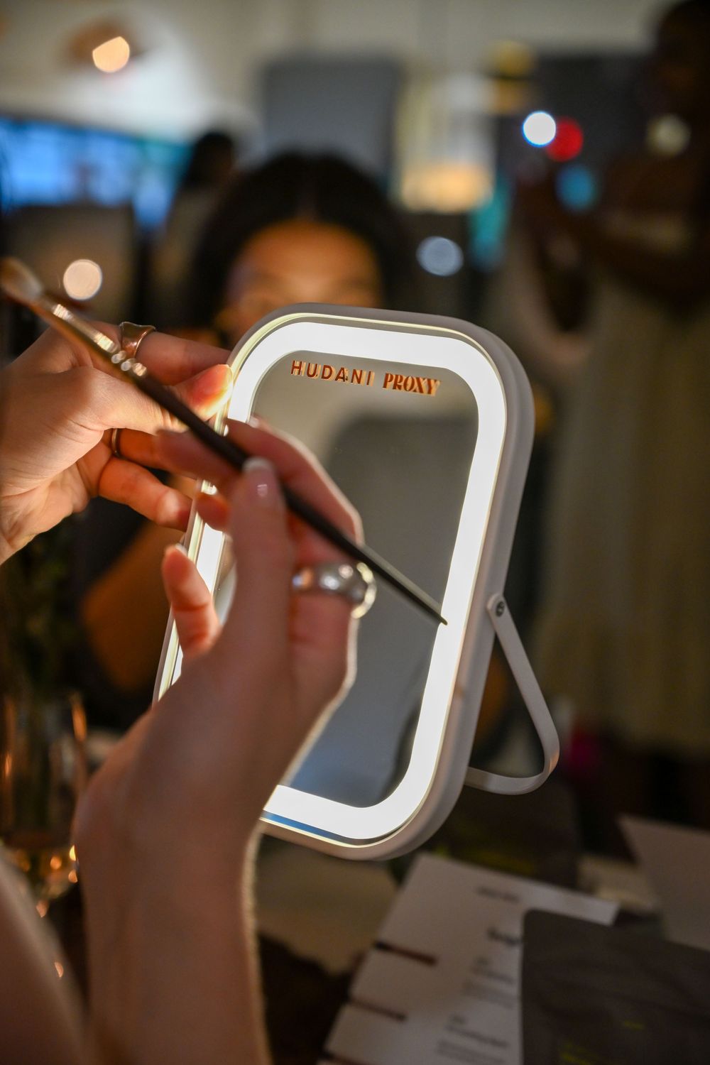 Hands holding makeup brushes near a lit Hudani Priay vanity mirror at a beauty event.