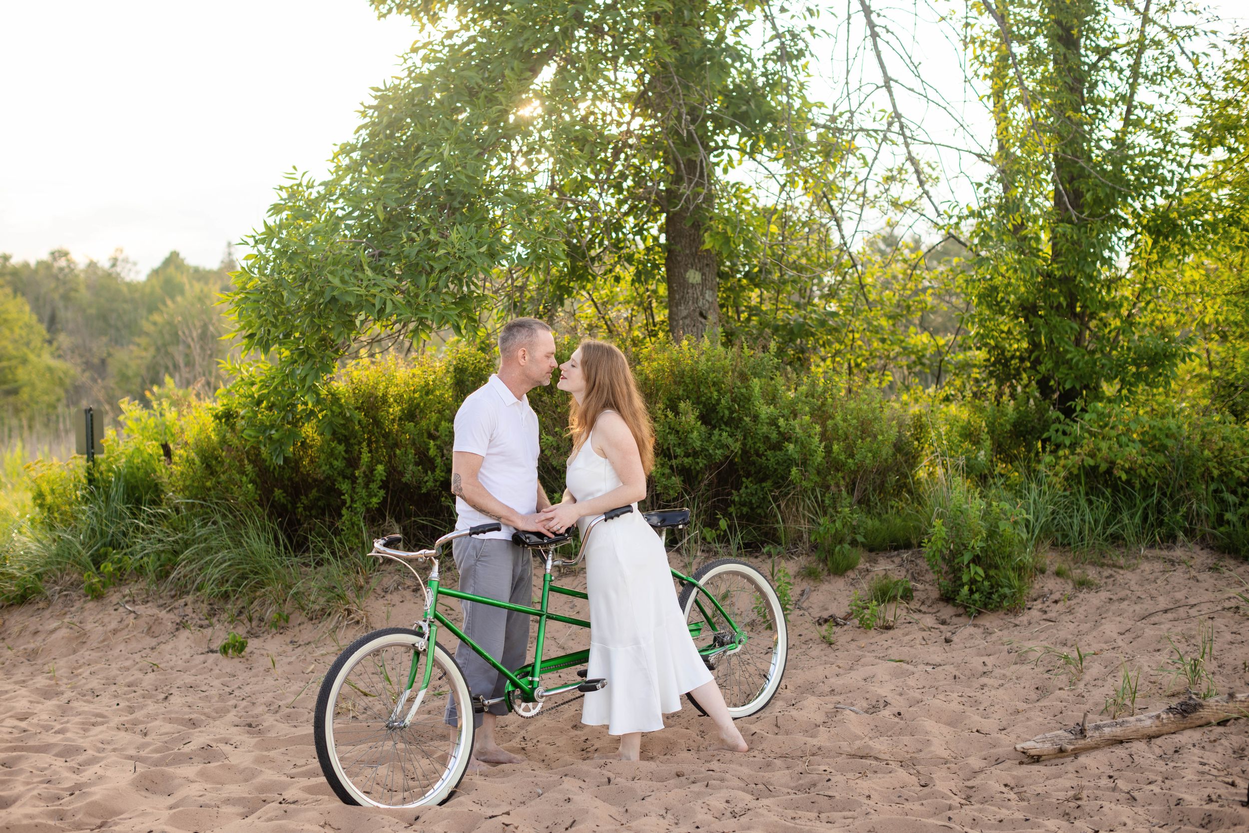 Couple holding hands beside a green tandem bicycle on sandy ground surrounded by lush green trees in golden sunlight.